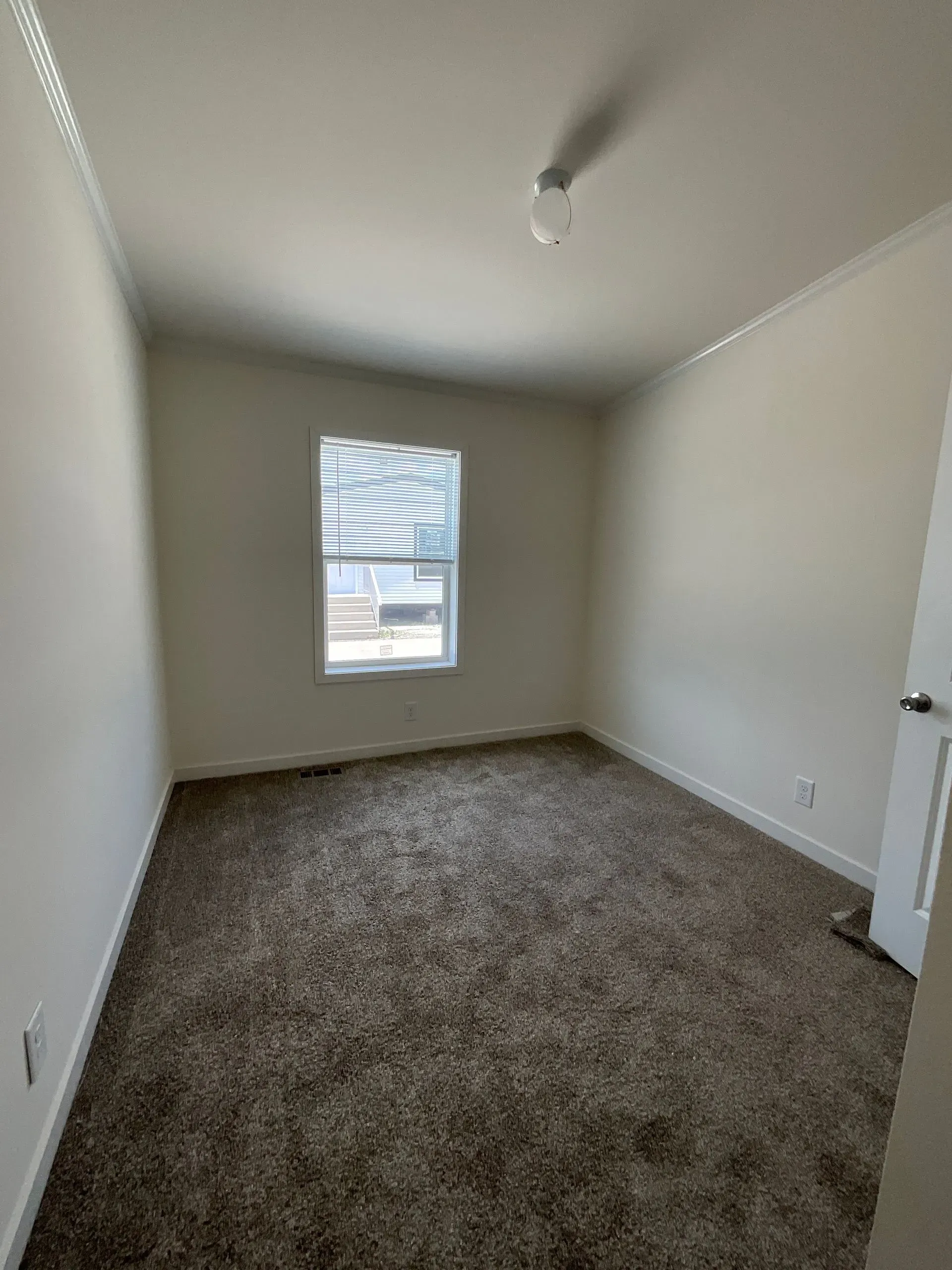 Empty room with brown carpet, window, and white walls. A bare lightbulb hangs from the ceiling.