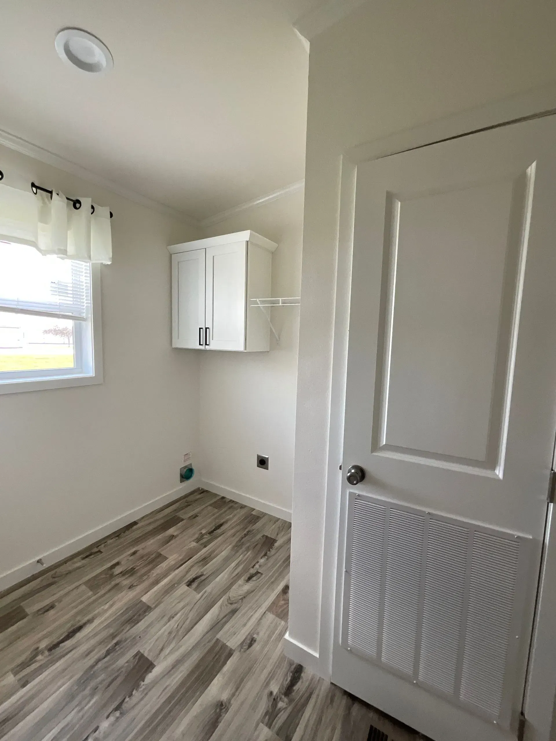 A small, white-walled room with a window, cabinet, and closed door. Wood-look flooring.