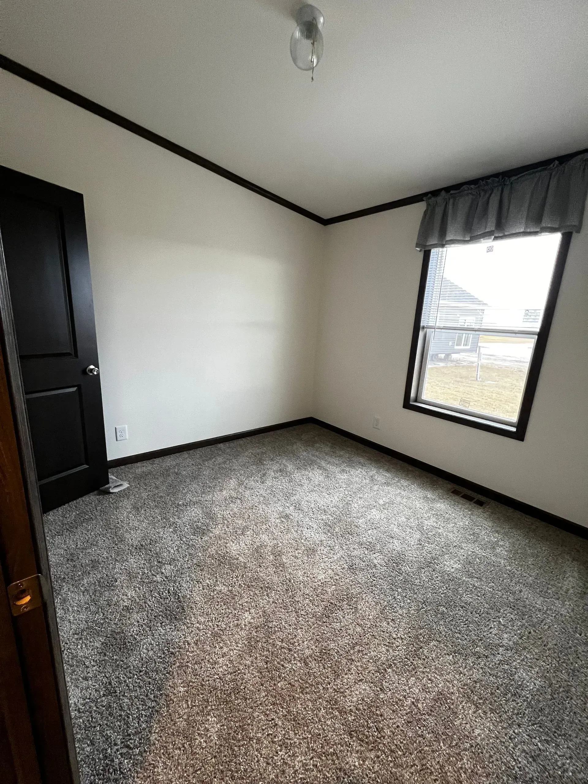 Empty bedroom with gray carpet, black trim, and a window with a gray curtain. A black door is visible.