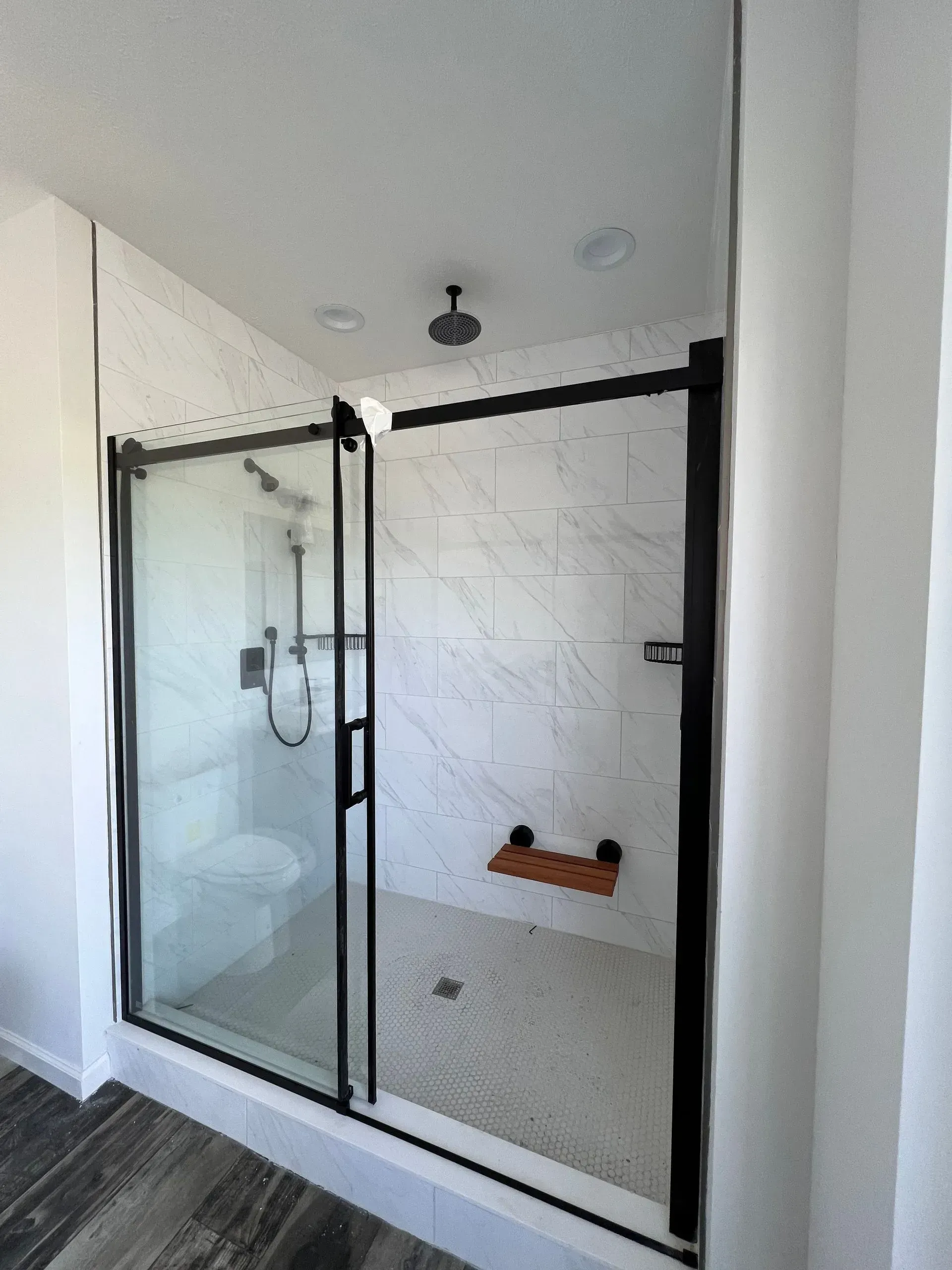 Modern shower with black-framed glass doors, white tiled walls, pebble floor, and wooden seat.