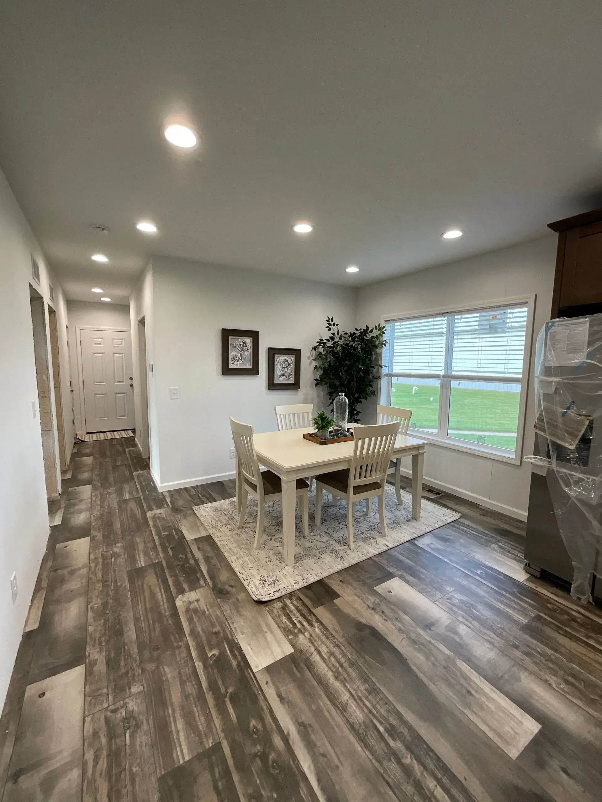 Dining room with white table, chairs, rug, and plant near a window. Wooden floors.