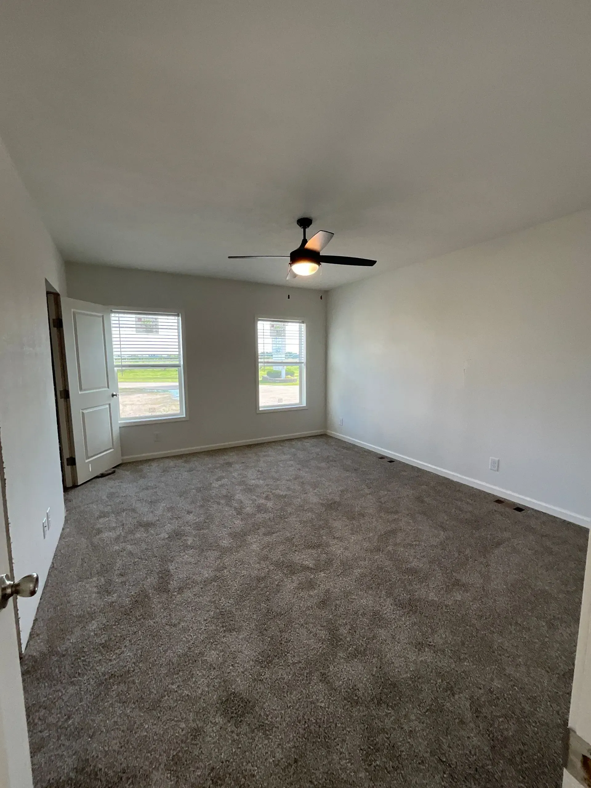 Empty room with gray carpet, white walls, two windows, and a ceiling fan.