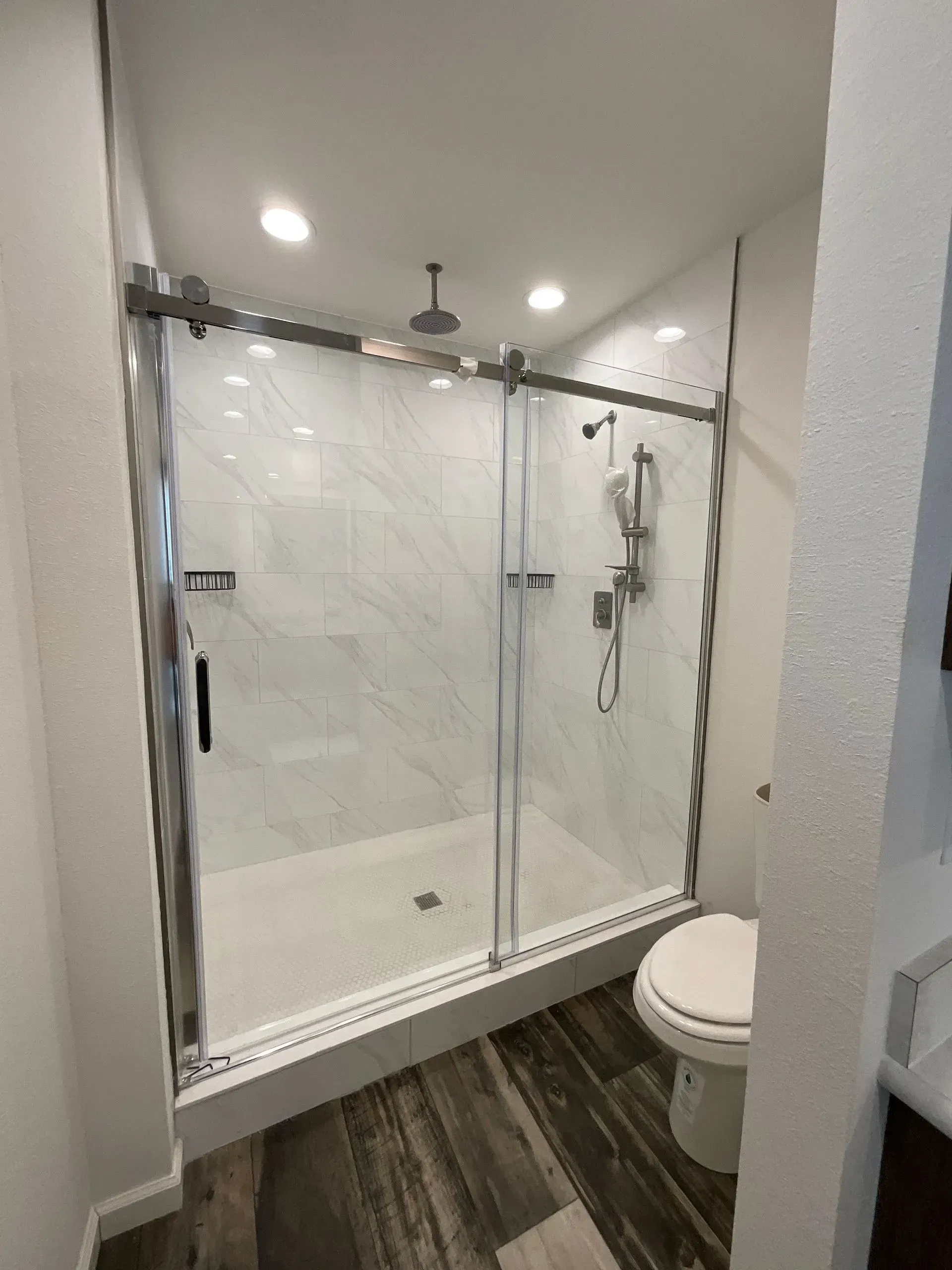 Shower with sliding glass doors, white tiled walls, and a toilet. The floor is wood-look tile.