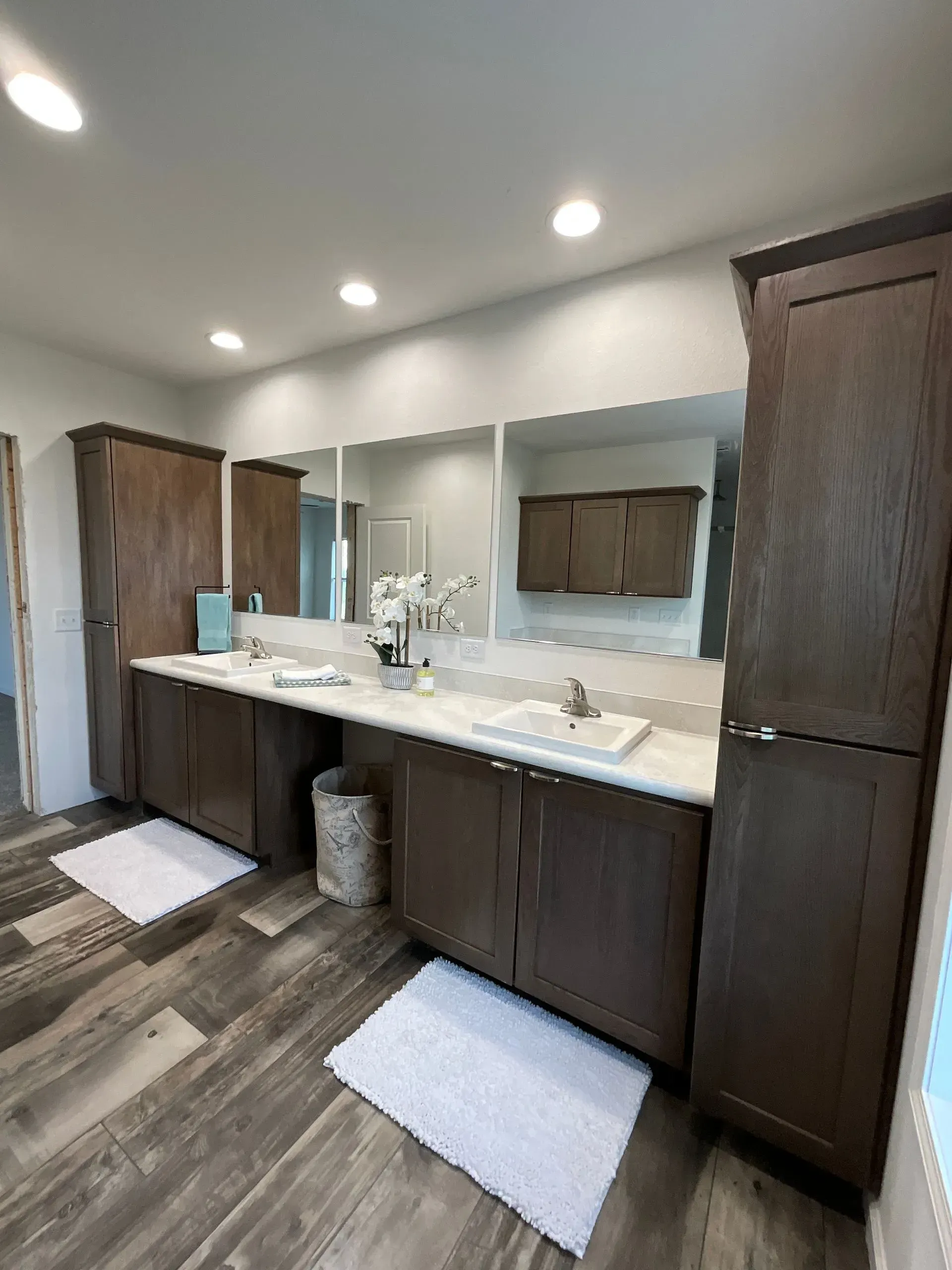 Bathroom with brown cabinets, long countertop with sinks, and white rugs on the floor.