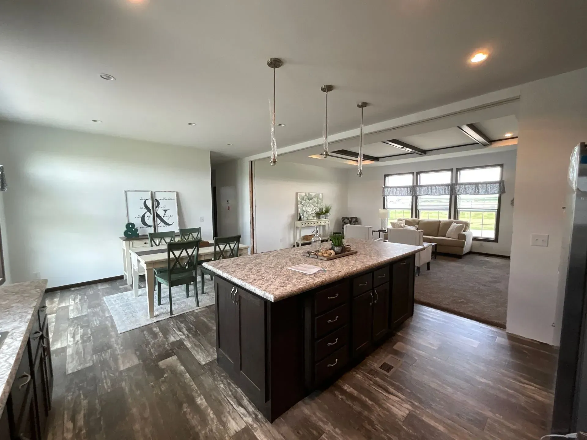 Open-concept kitchen with dark brown island and dining area, leading to a living room with large windows.