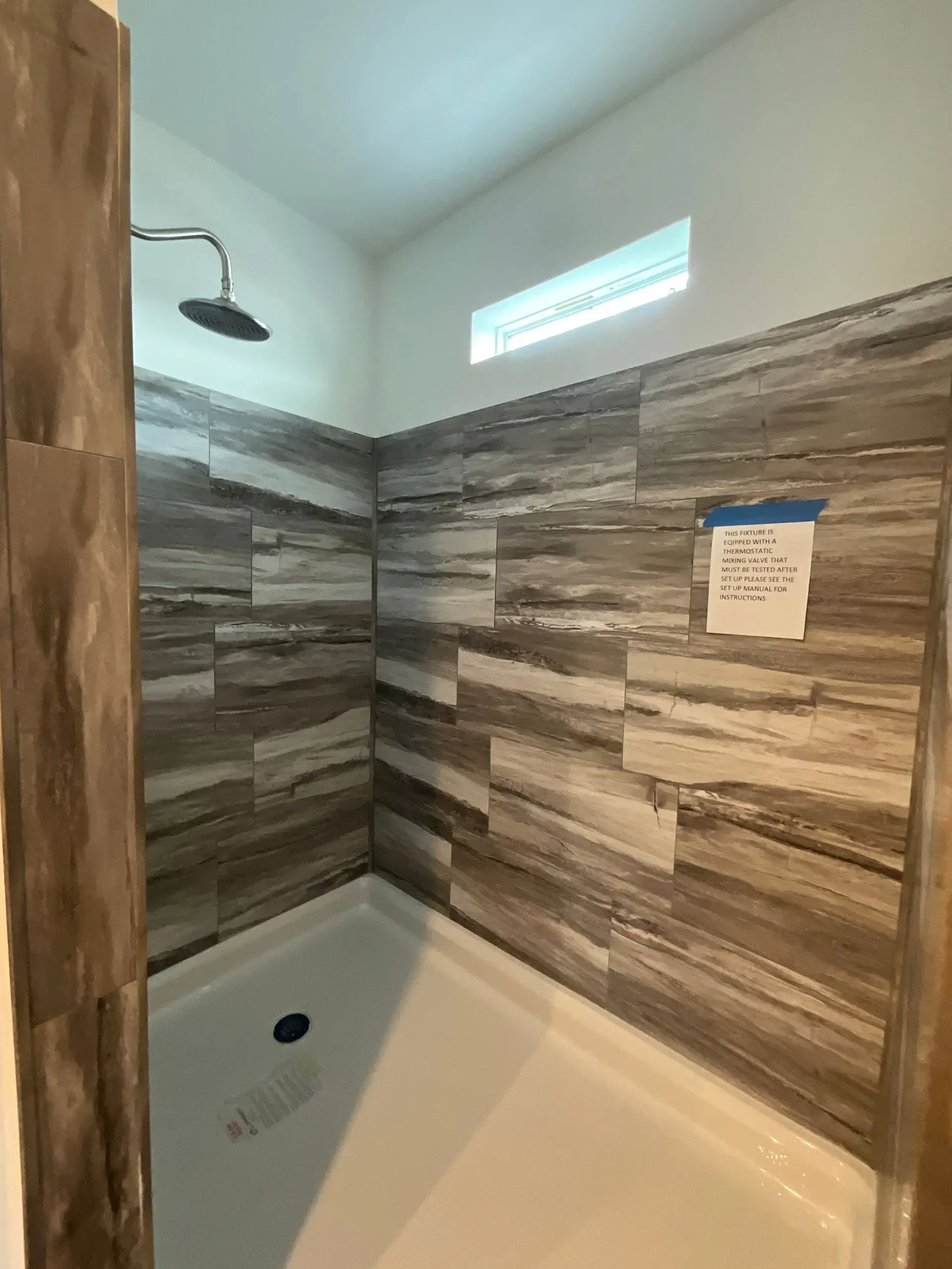 Shower stall with gray and brown tiled walls, a white shower pan, and a single shower head.