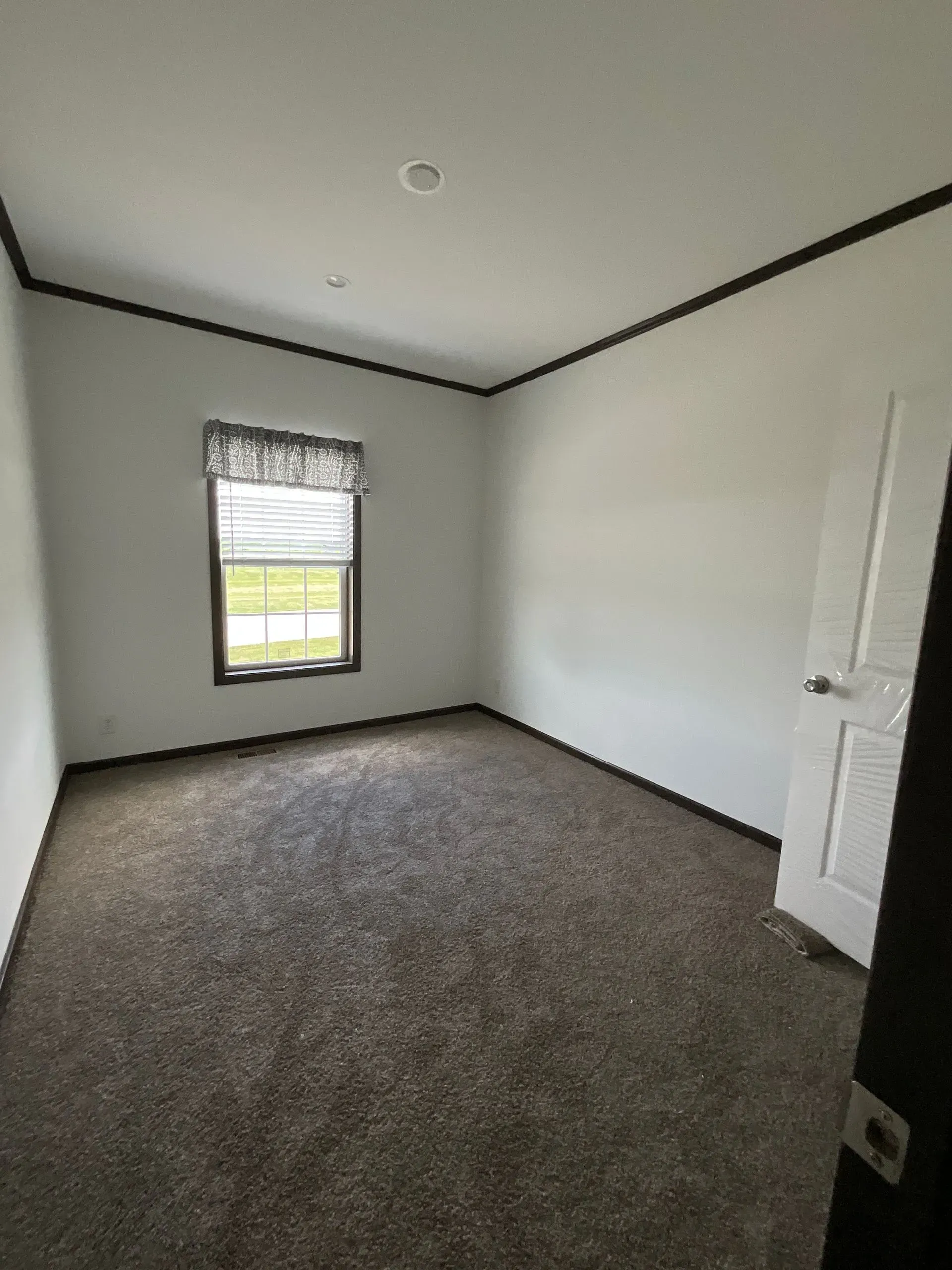 Empty room with brown carpet, white walls, dark trim, a window with blinds, and a white door.