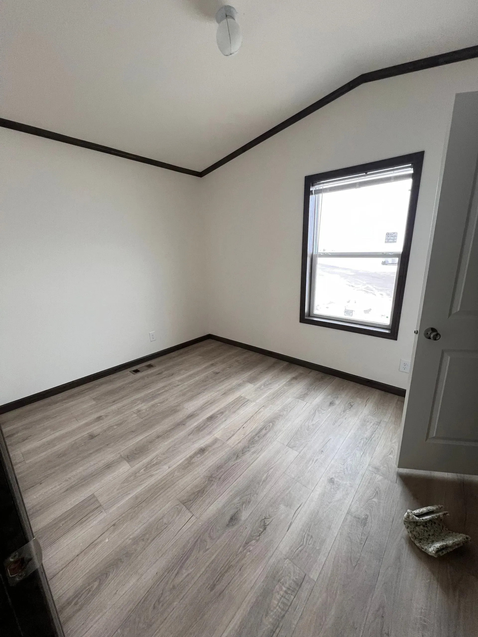 Empty room with light wood-look flooring, white walls, black trim, window, and closed door.