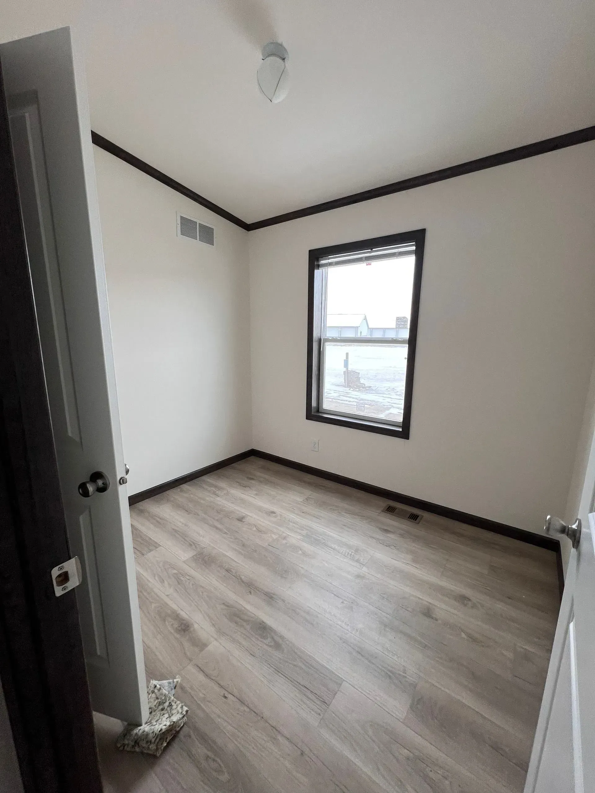 Empty room with light wood floor, white walls, dark trim, and a window. Doorway on the left.