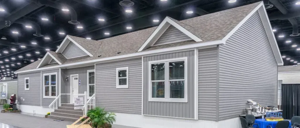 A gray modular home on display in an event hall. It has a gabled roof, white trim and several windows.