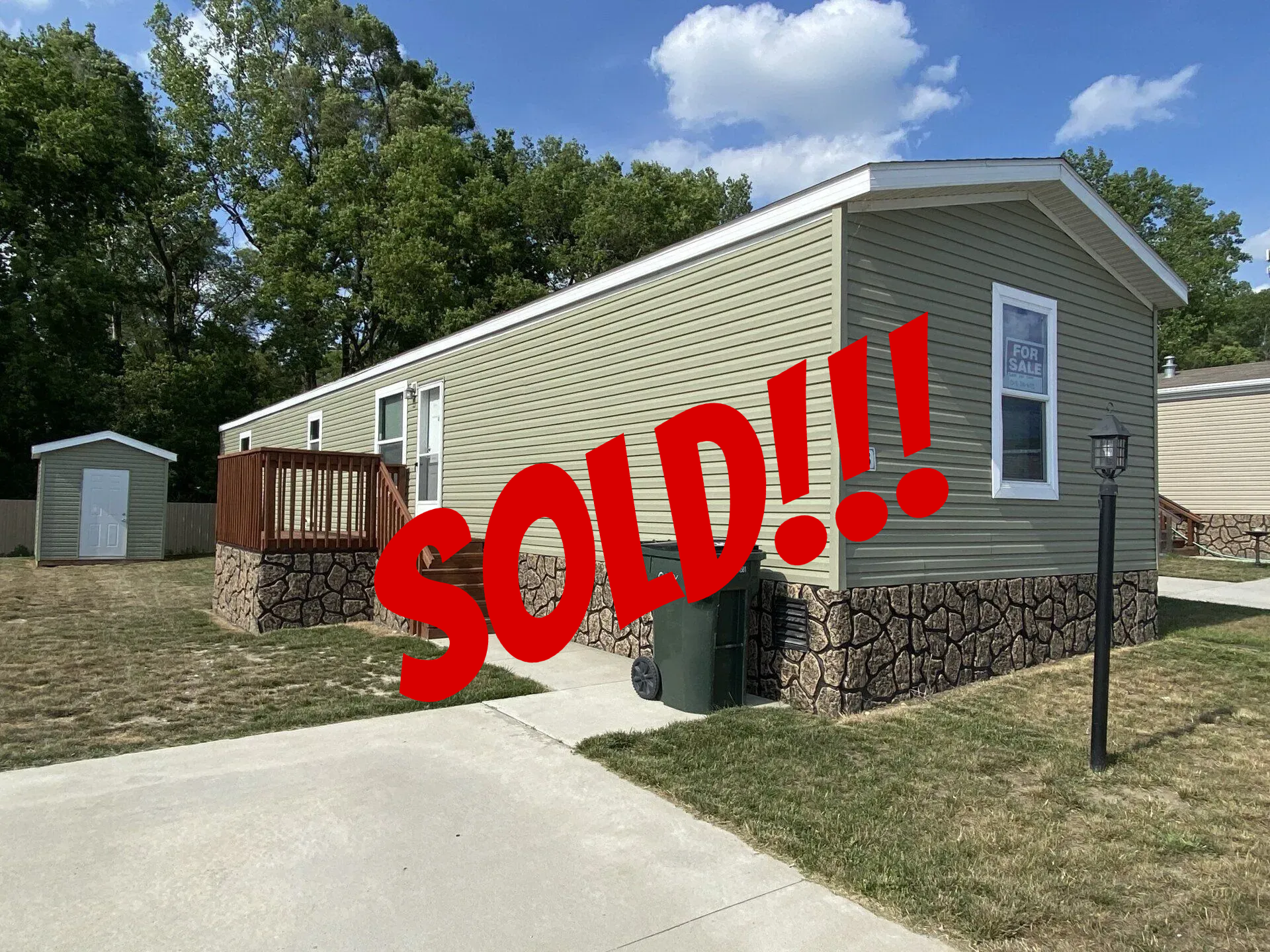 Green mobile home with stone foundation and small shed, sunny day.