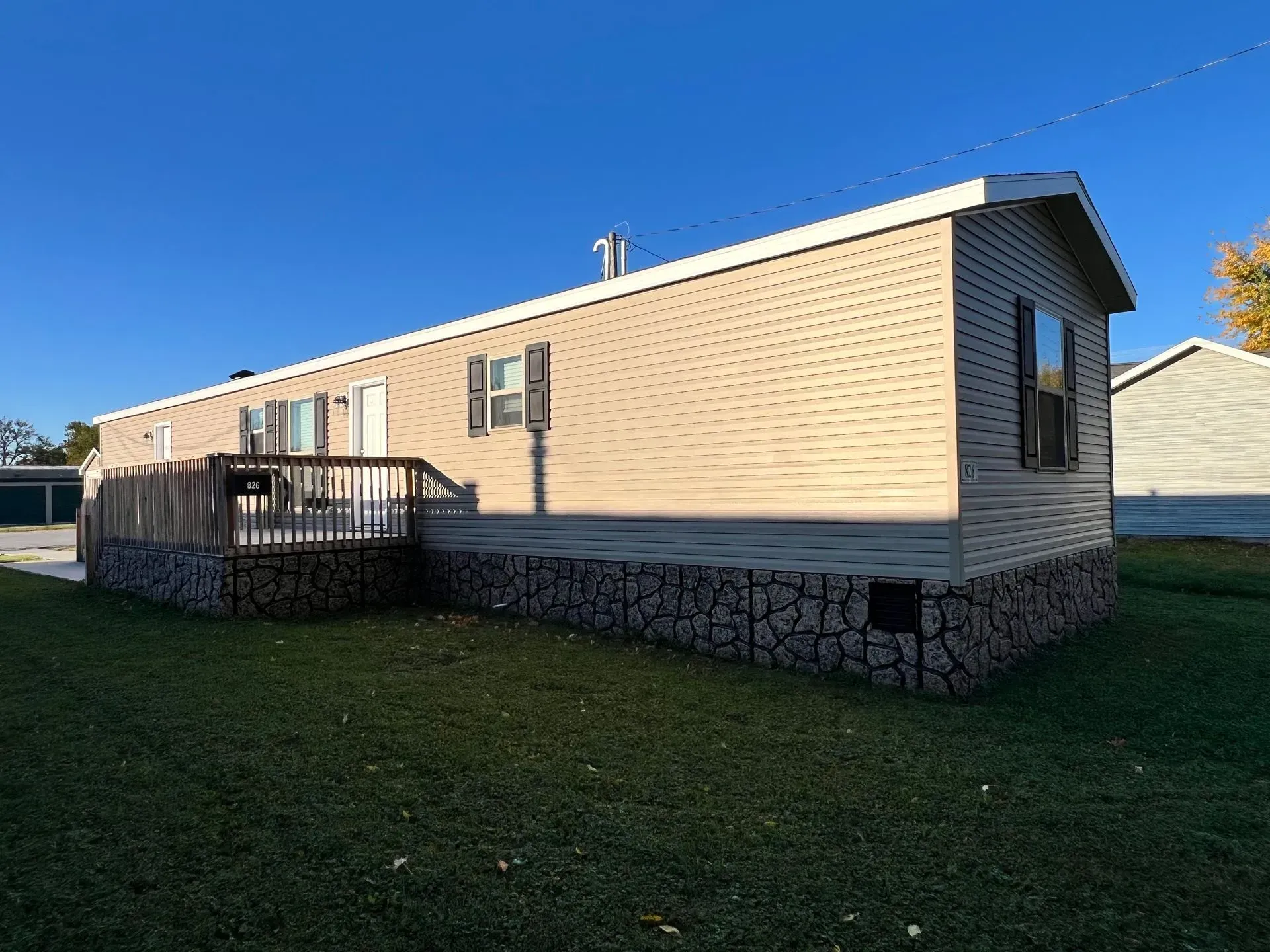 Tan mobile home with brown shutters, a deck, and rock skirting under a blue sky.