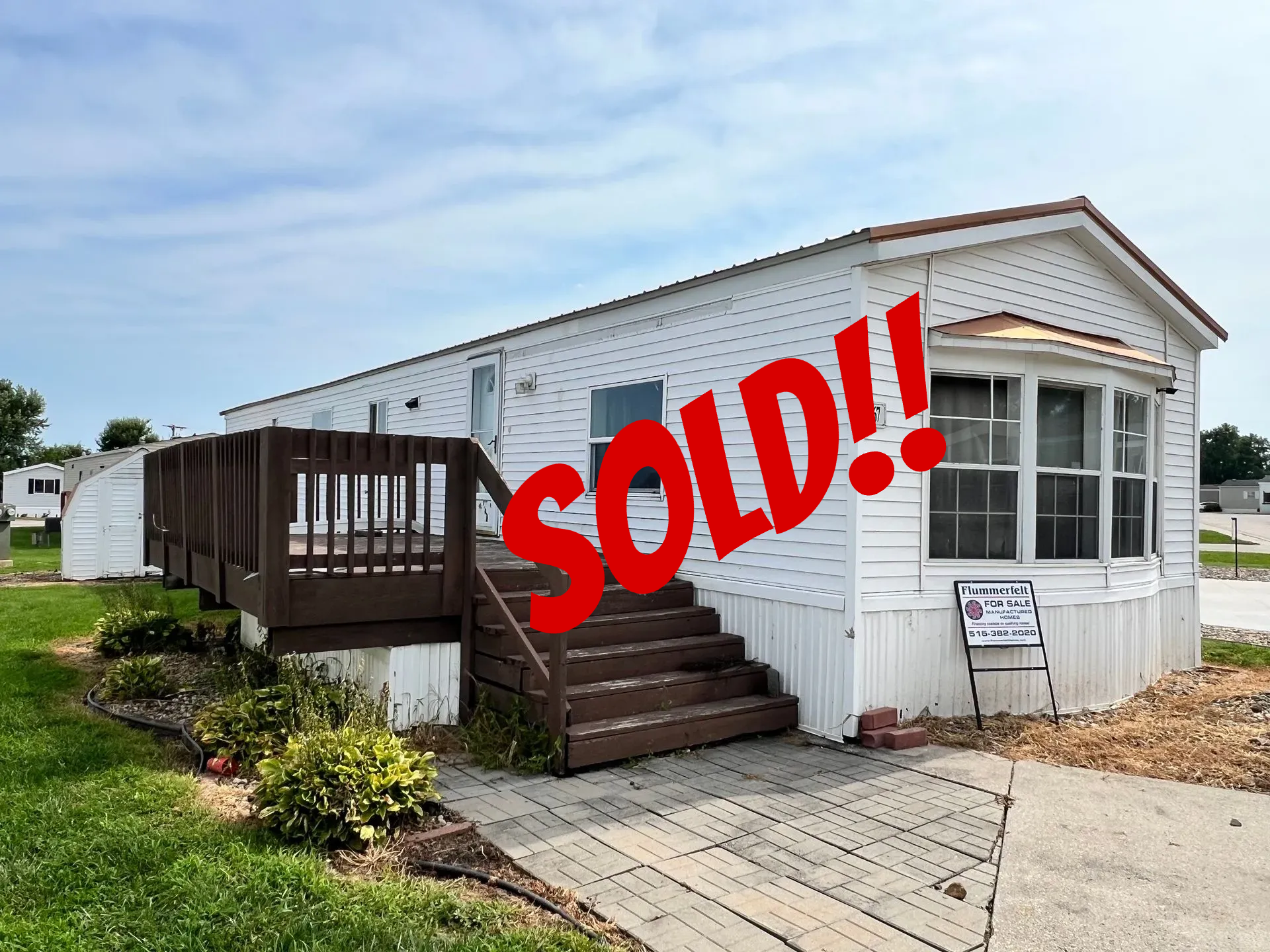 White mobile home with brown deck and stairs.  Gray sidewalk leads up to the home with shrubs and a for sale sign.