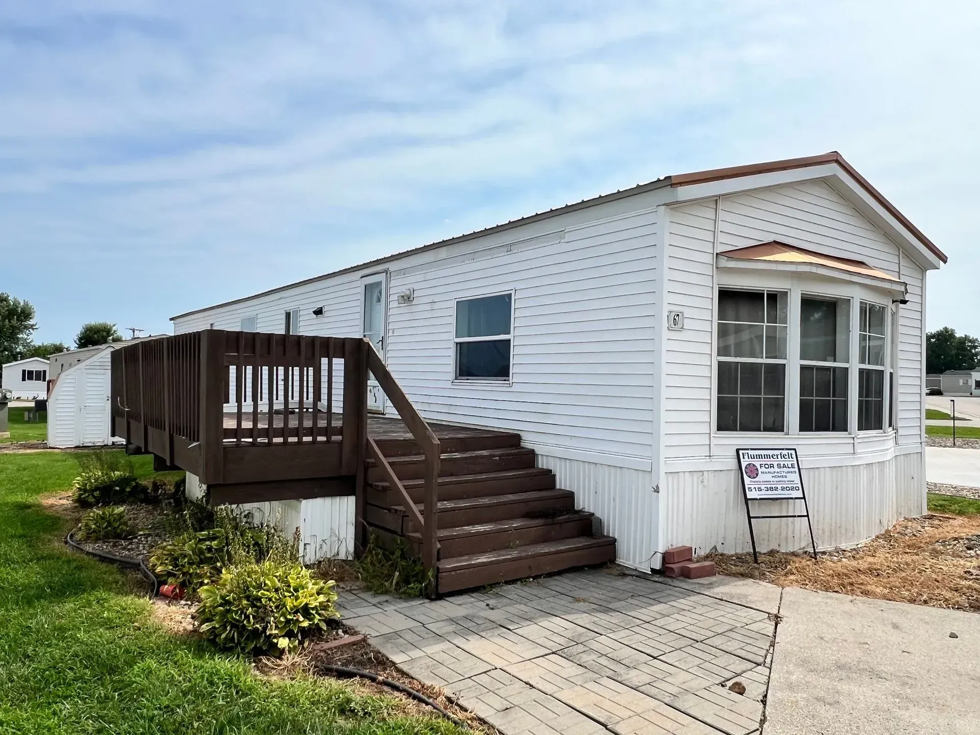 White mobile home with brown deck and stairs. Gray sidewalk leads up to the home with shrubs and a for sale sign.