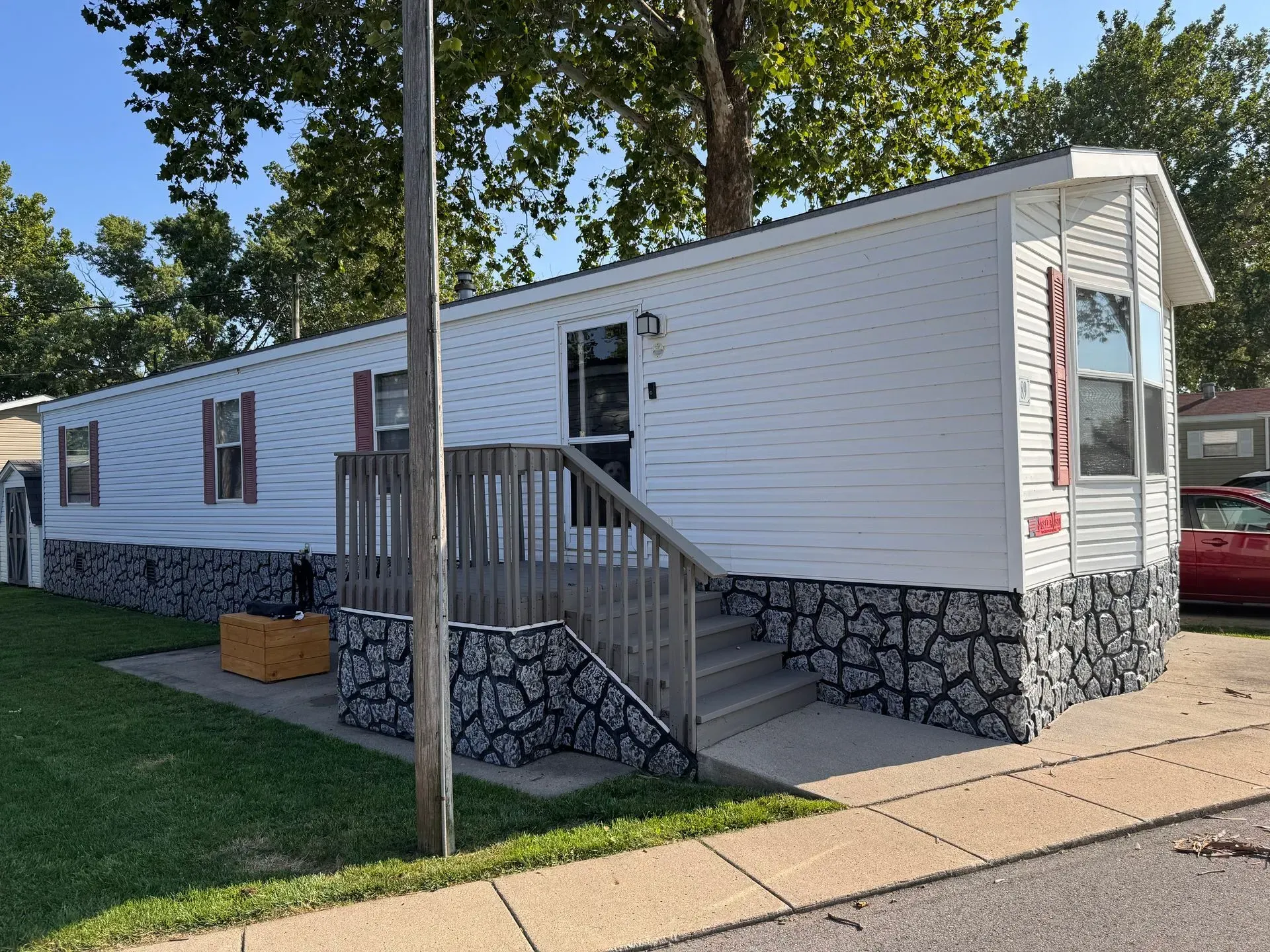 Mobile home with white siding, stone-patterned base, red shutters, and wooden stairs.