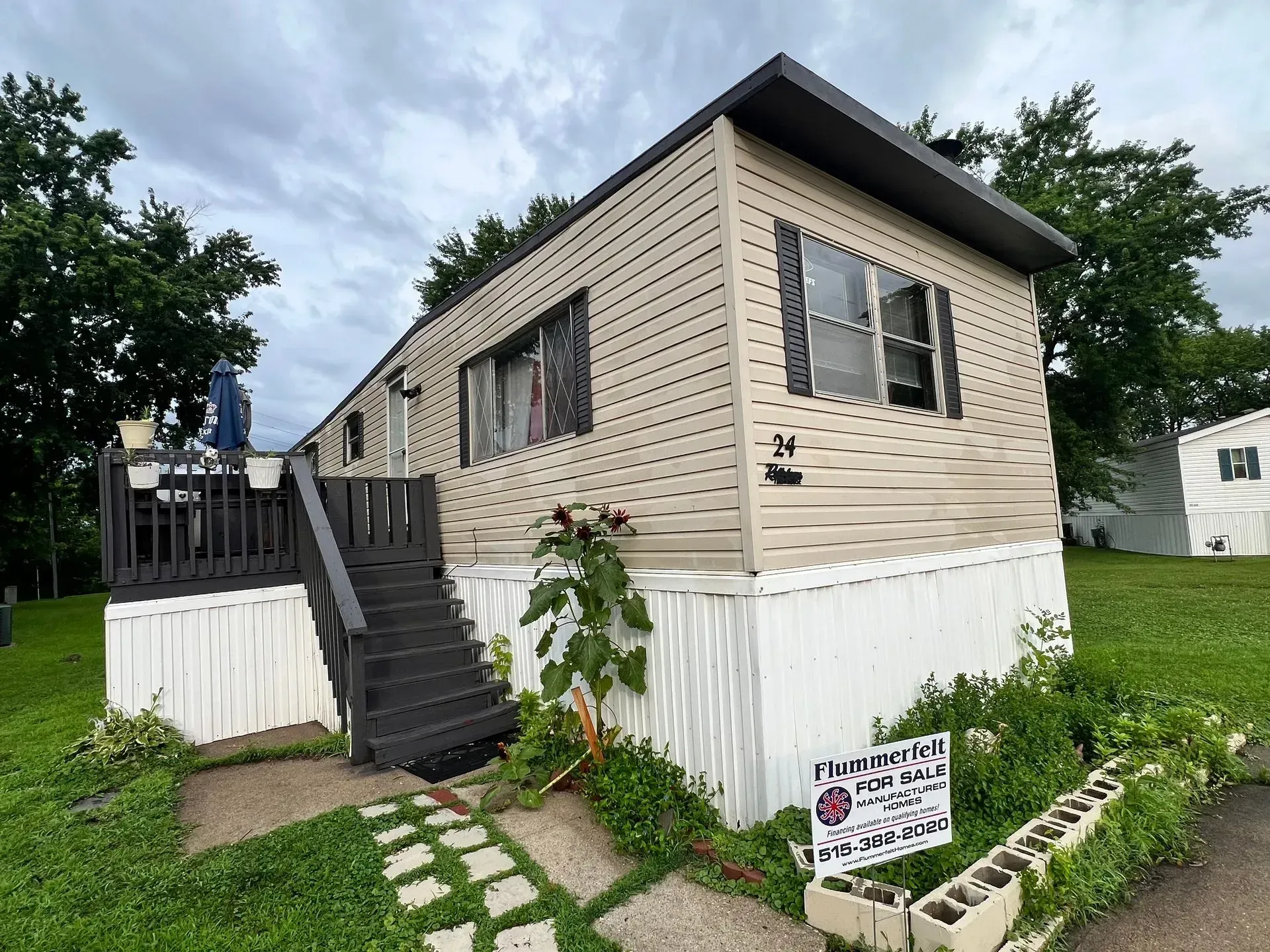 Mobile home with a deck and steps, surrounded by grass and plants, under a cloudy sky.