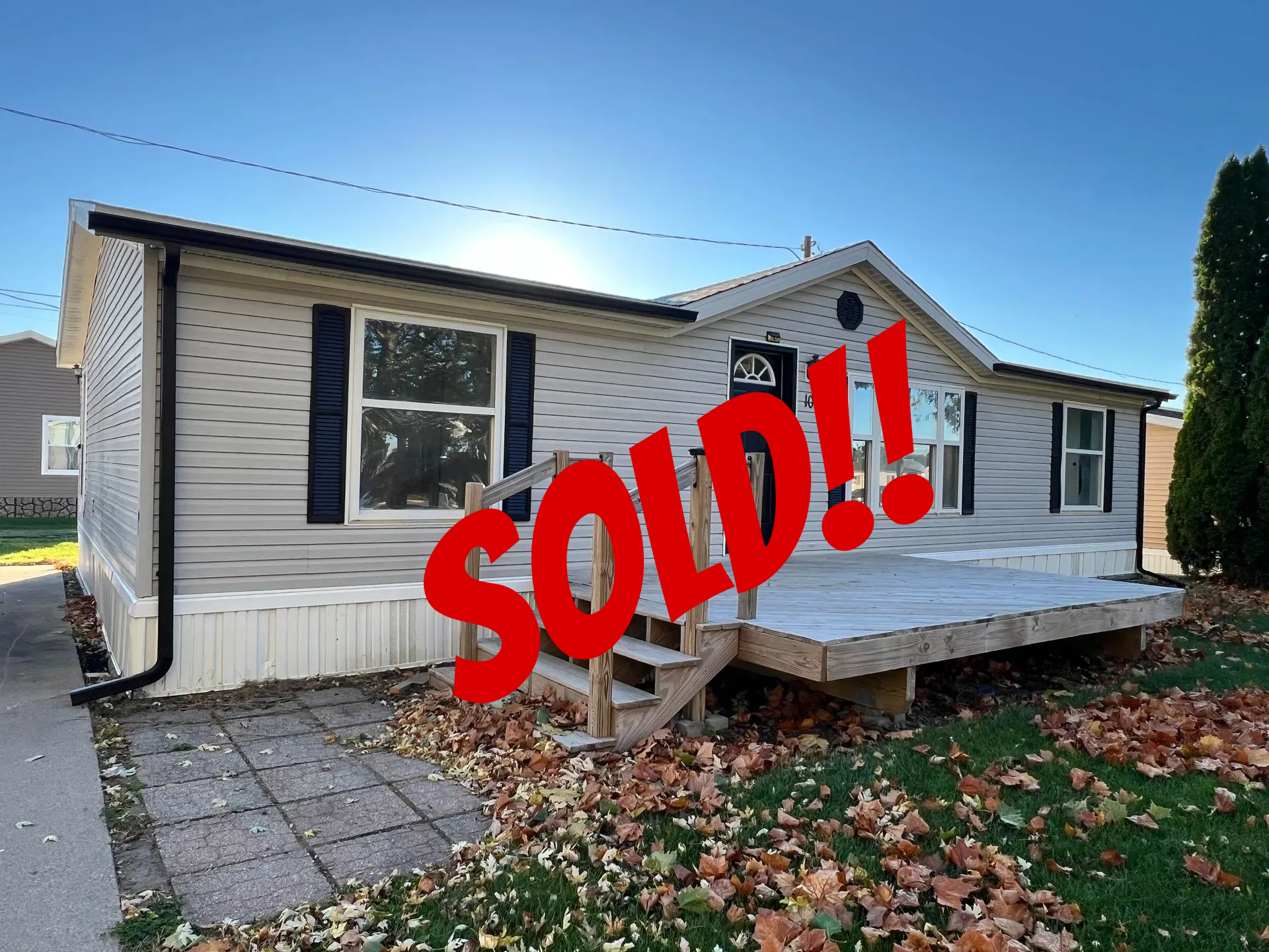 Gray-sided mobile home with black shutters, wooden deck, and steps; autumn leaves on the lawn.