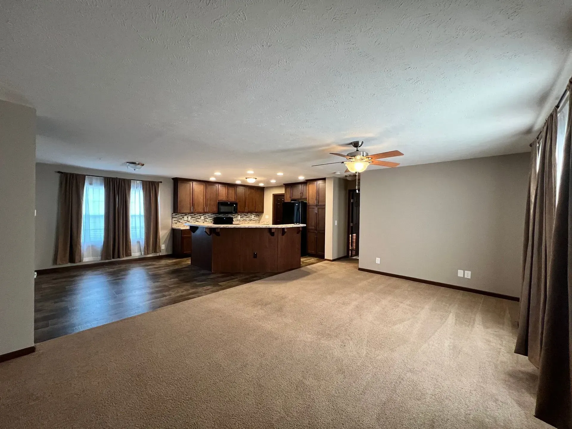 Open-plan living space with kitchen island, brown cabinetry, carpet, hardwood floor, and a ceiling fan.