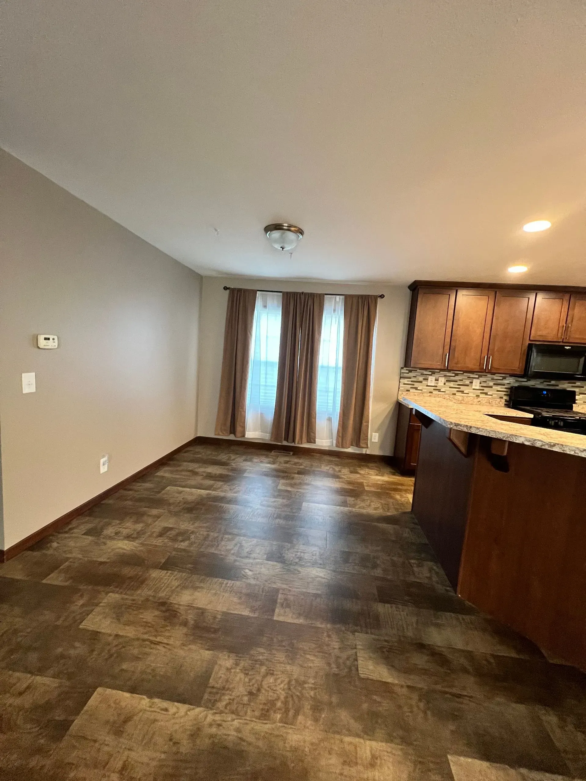 Empty living space with brown flooring, tan walls, kitchen cabinets, and curtains.