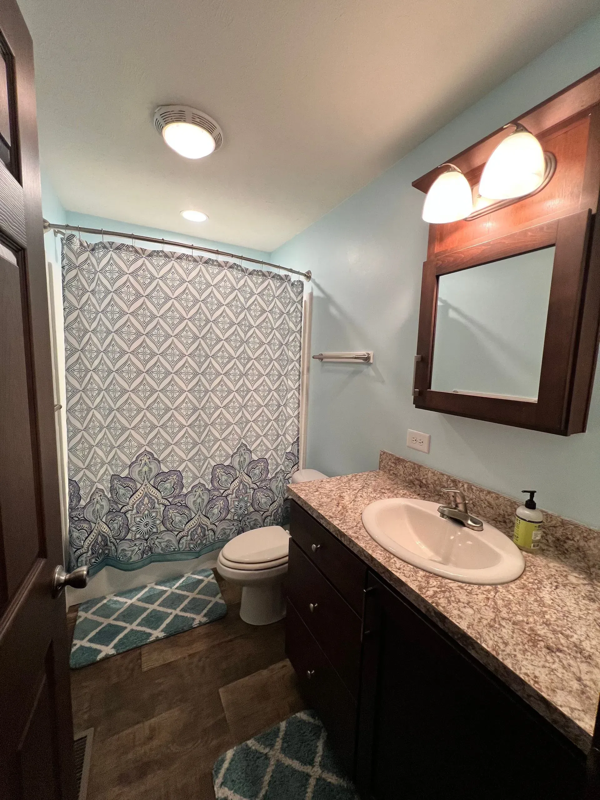 Bathroom with blue walls, dark brown vanity and door, shower curtain, and teal rugs.