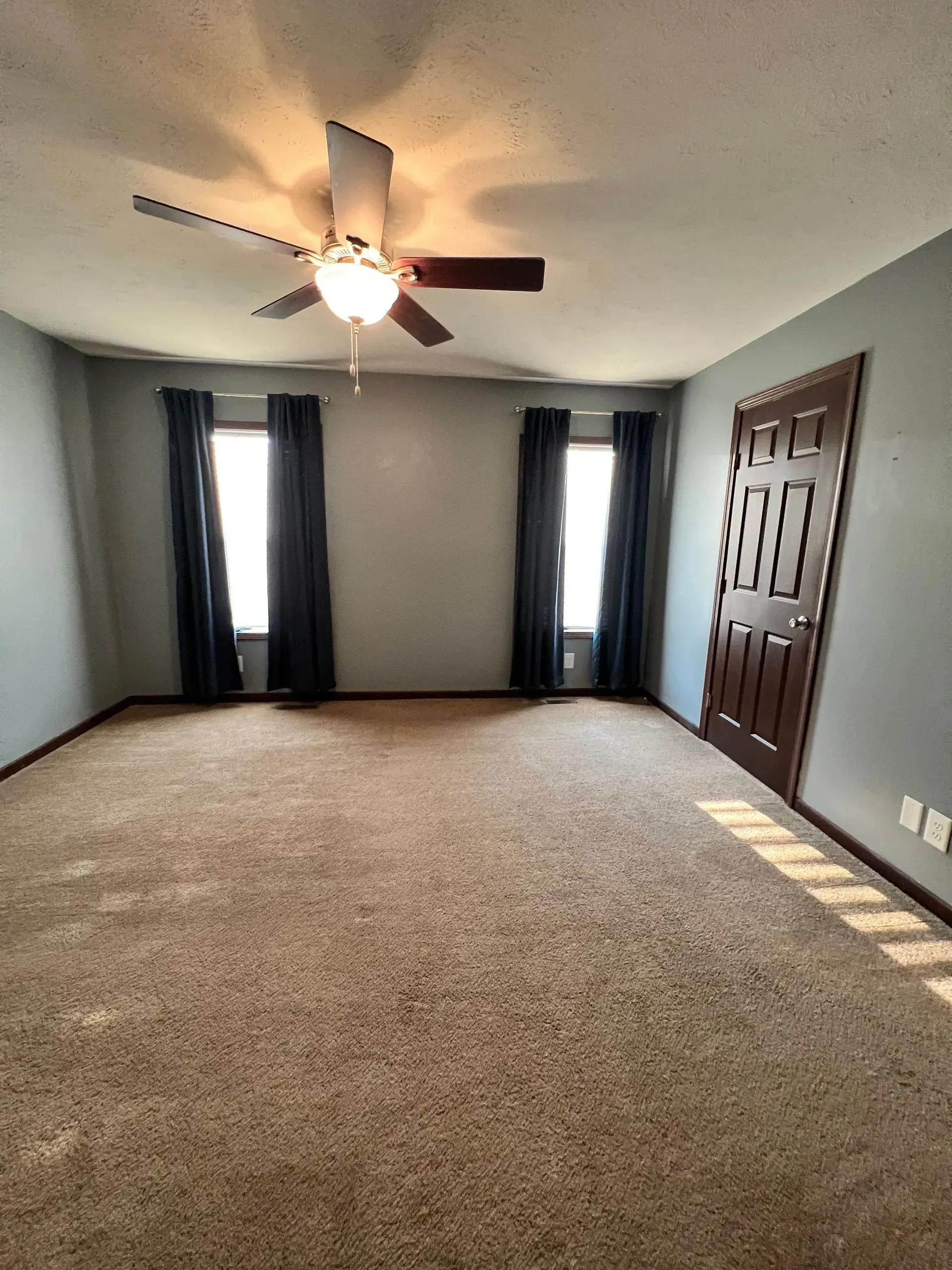 Empty room with brown carpet, two windows with black curtains, and a brown door.
