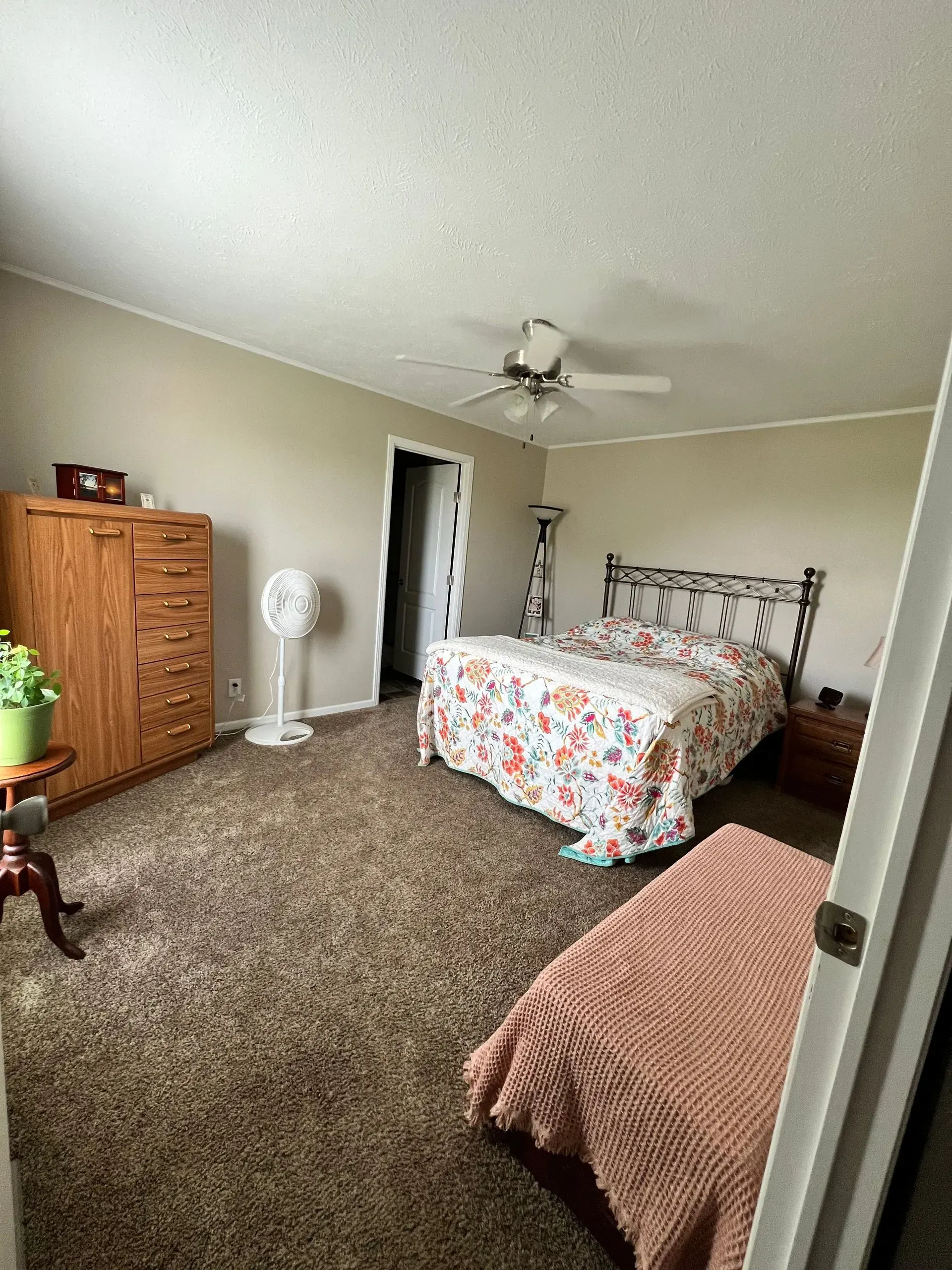 Bedroom with a bed, dresser, fan, and an open doorway. Brown carpet, tan walls, and a floral bedspread.