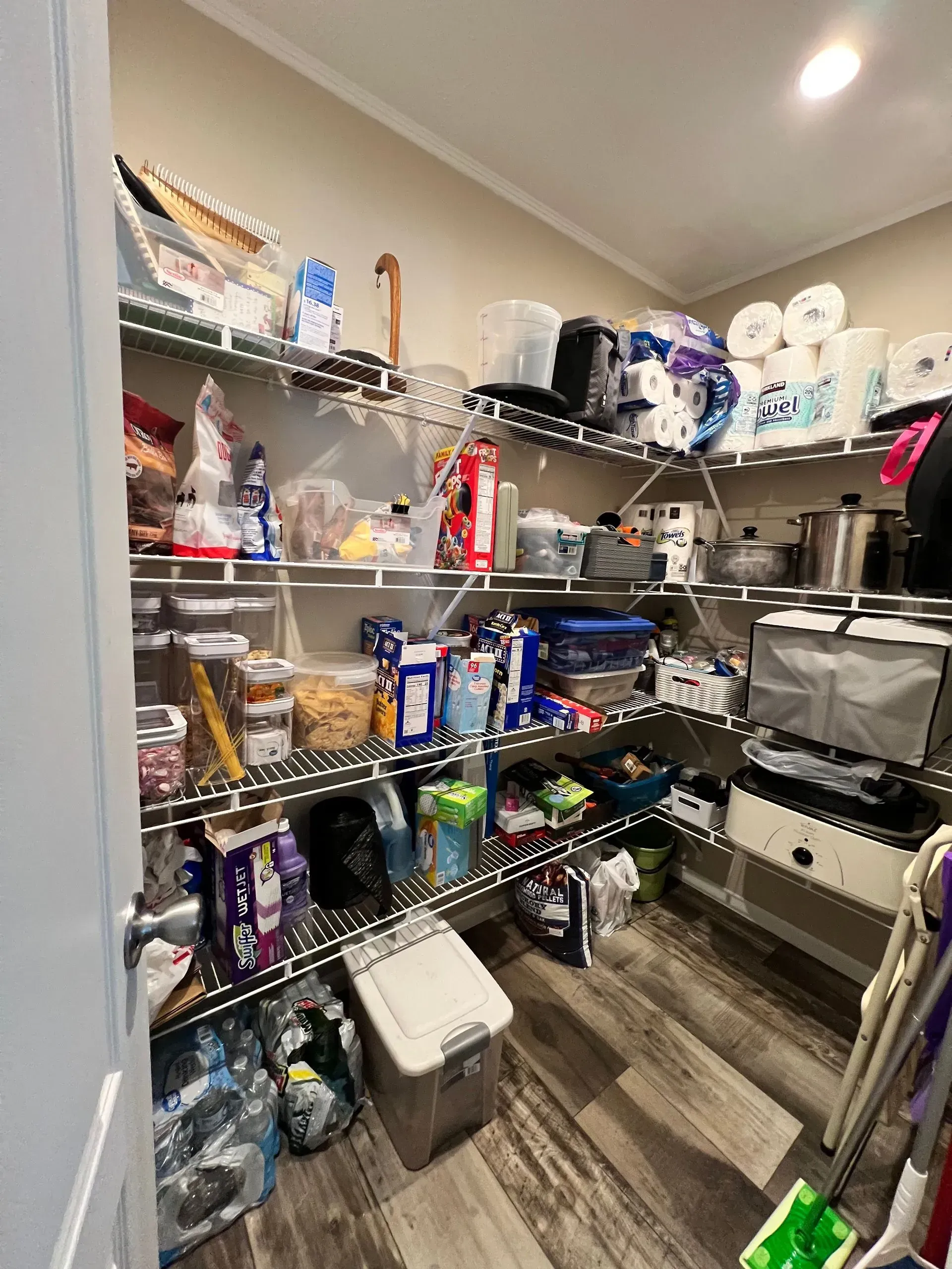 Cluttered pantry with wire shelving, food items, and various containers. Wood-look flooring.