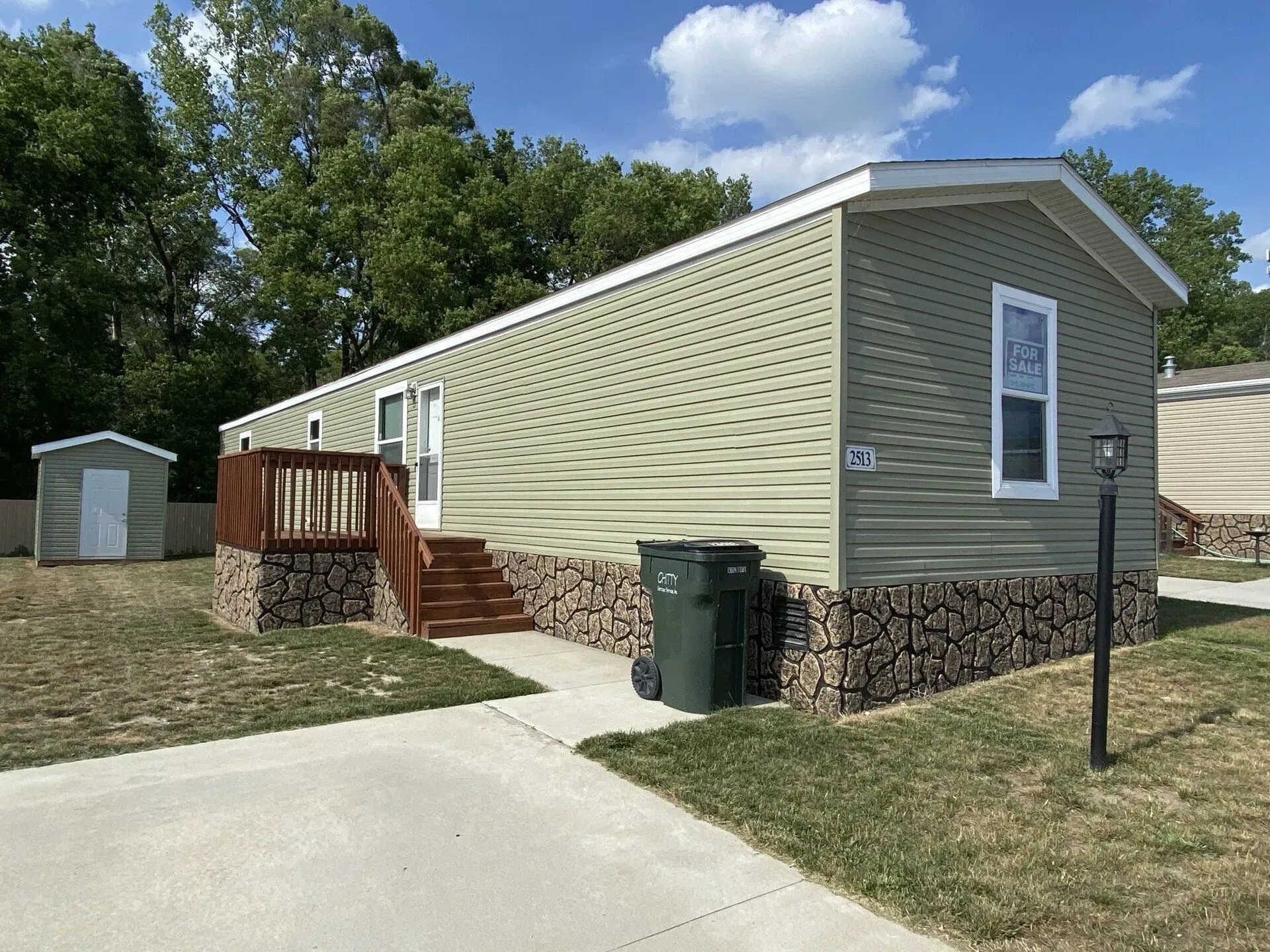 Green-sided mobile home with wood steps, stone base, and a small shed on a sunny day.