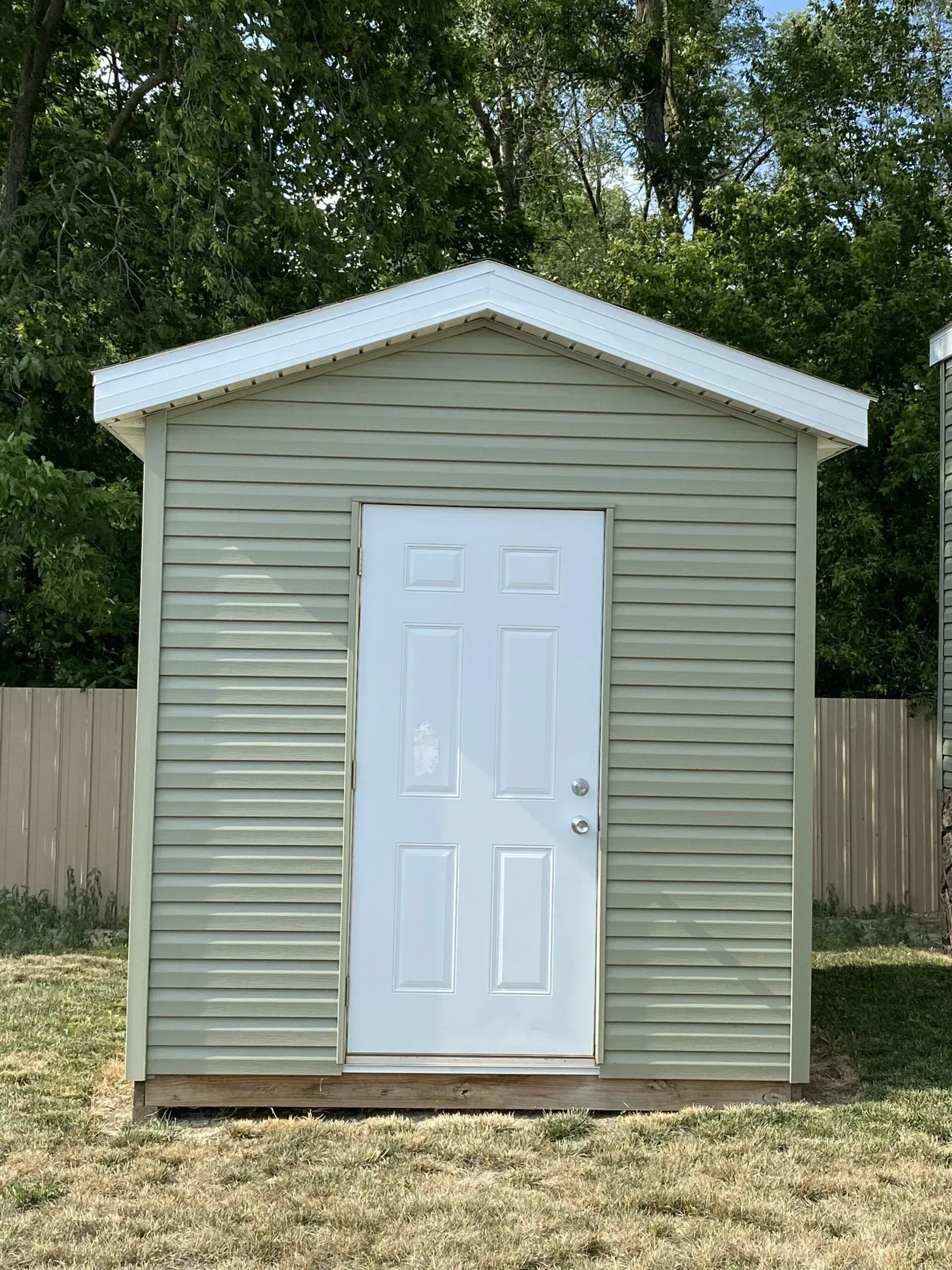 Green storage shed with white door, set in a grassy yard, with a wooden fence and trees in the background.