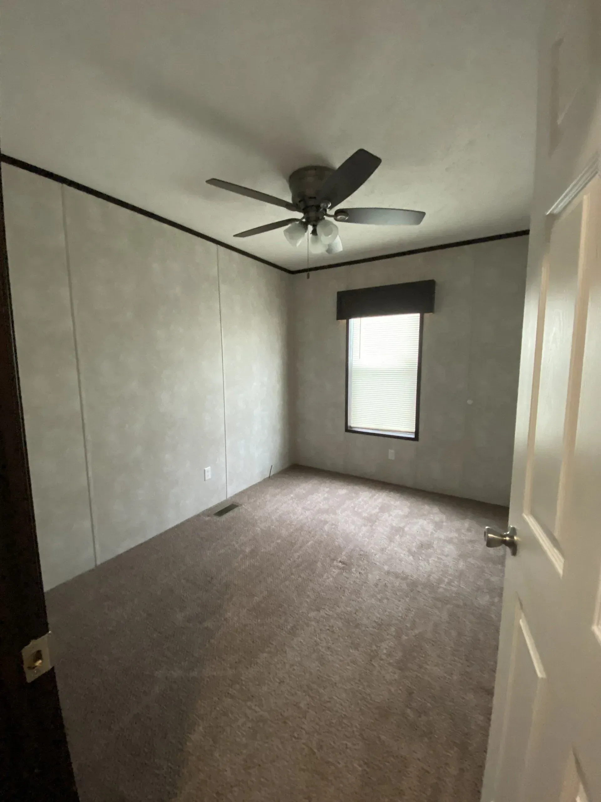 Empty bedroom with brown carpet, window, ceiling fan, and white door.