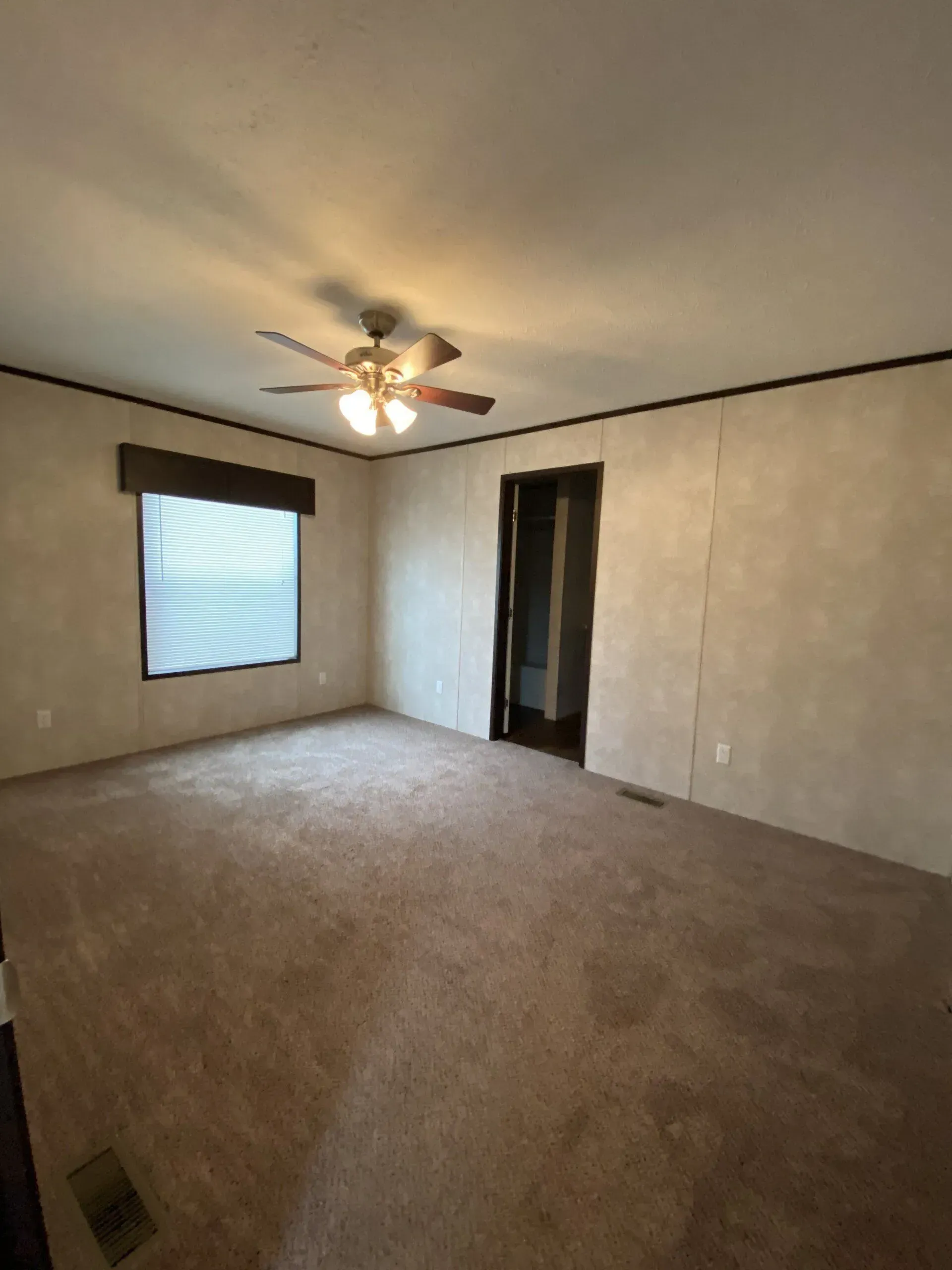 Empty bedroom with brown carpet, window with blinds, and a closet. Beige walls and a ceiling fan.
