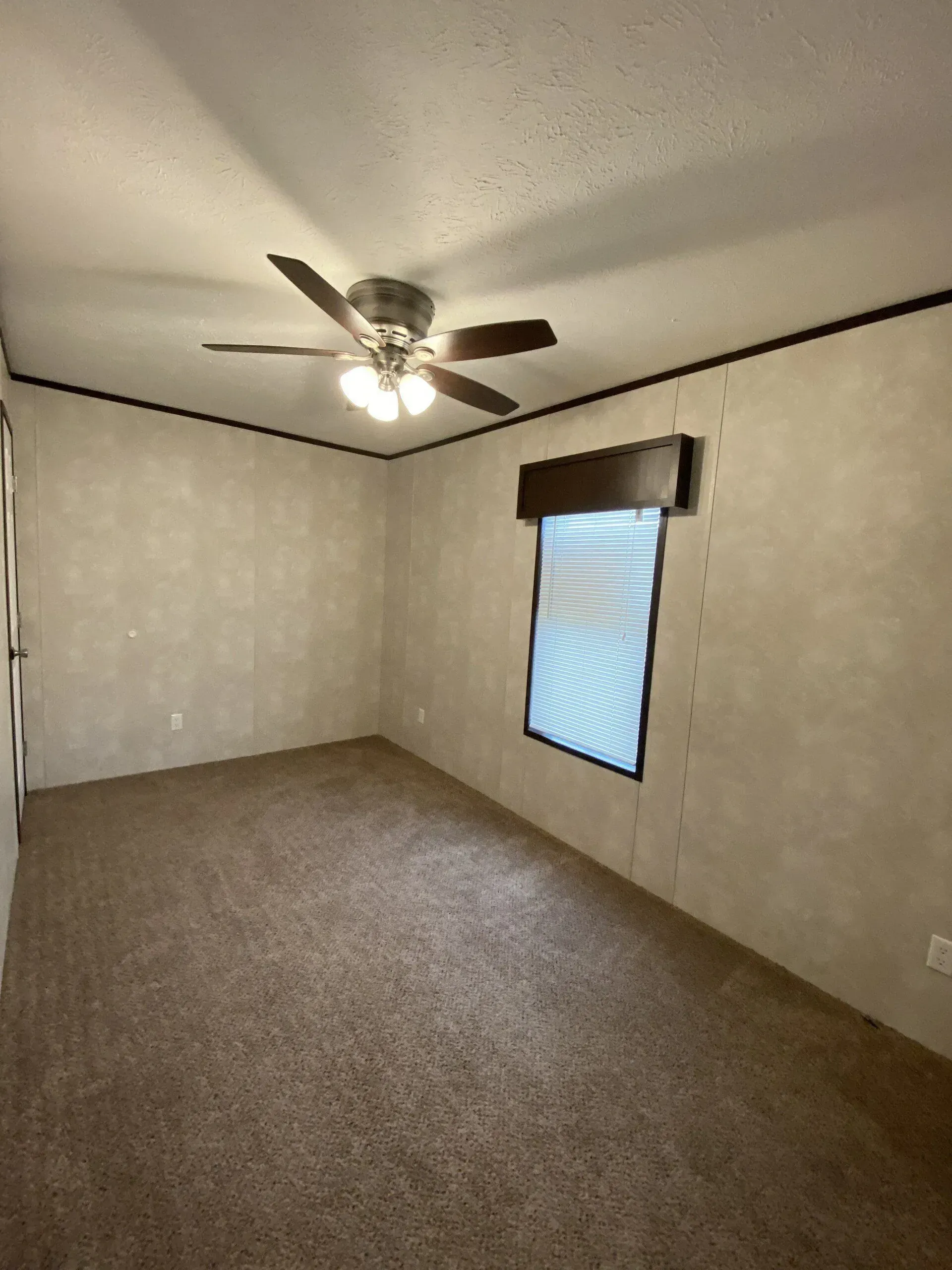 Empty bedroom with tan walls, brown carpet, ceiling fan, and a window.