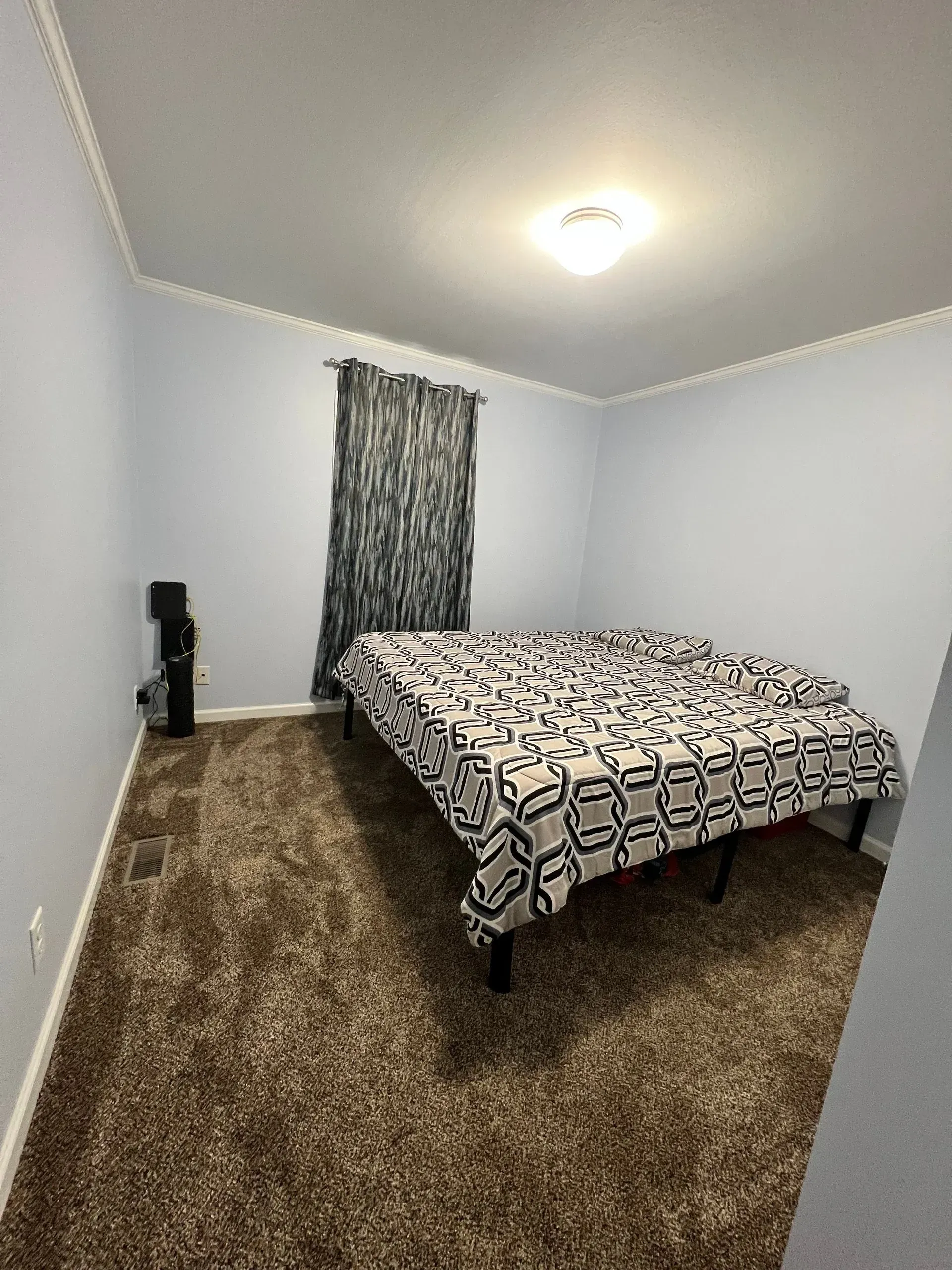 Bedroom with bed, patterned comforter, brown carpet, light blue walls, gray patterned curtains, and a black heater.