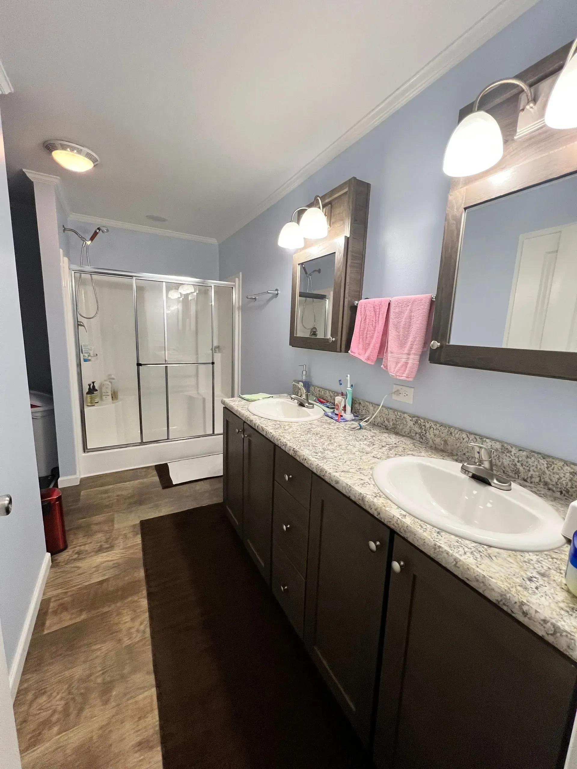 Bathroom with a double sink vanity, shower, brown cabinets, and light blue walls.