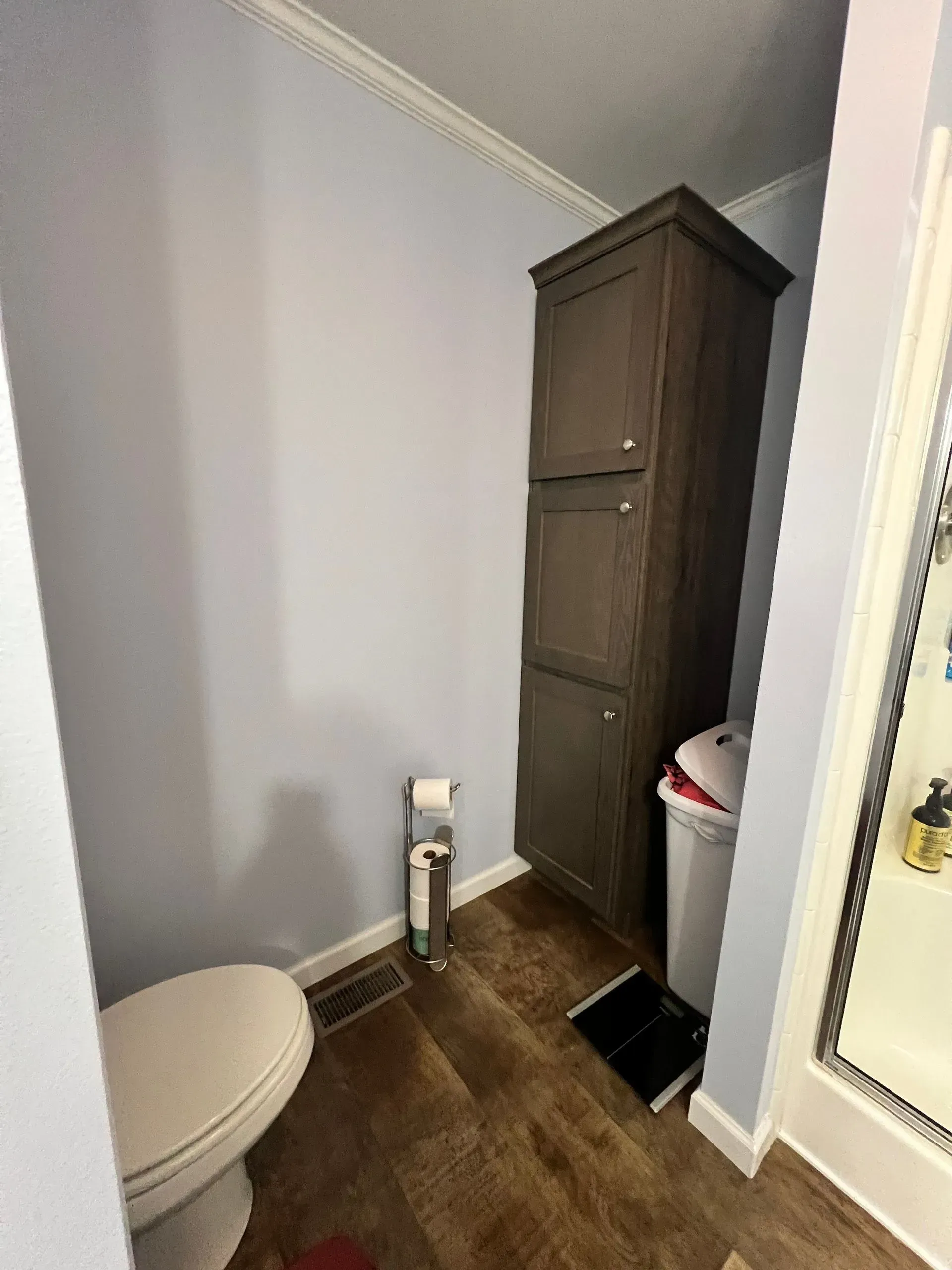 Bathroom interior with toilet, tall cabinet, and scale on a wood-look floor; light blue walls.