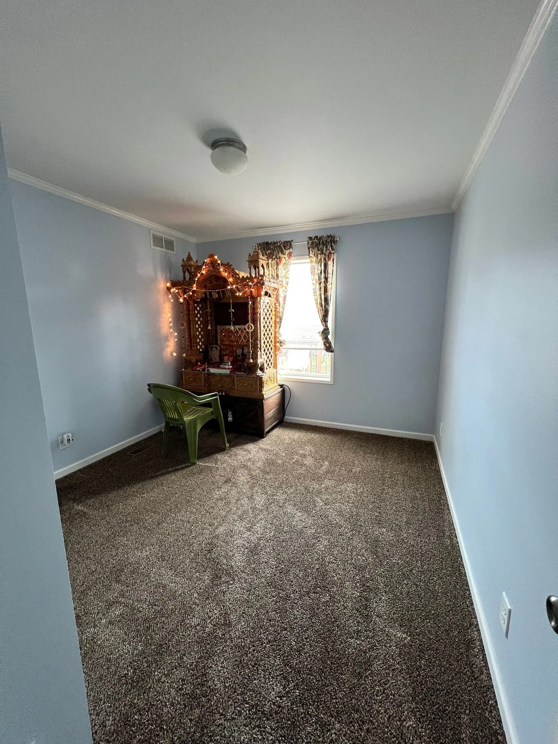 Blue-walled room with brown carpet, ornate wooden cabinet, chair, and small window with curtains.