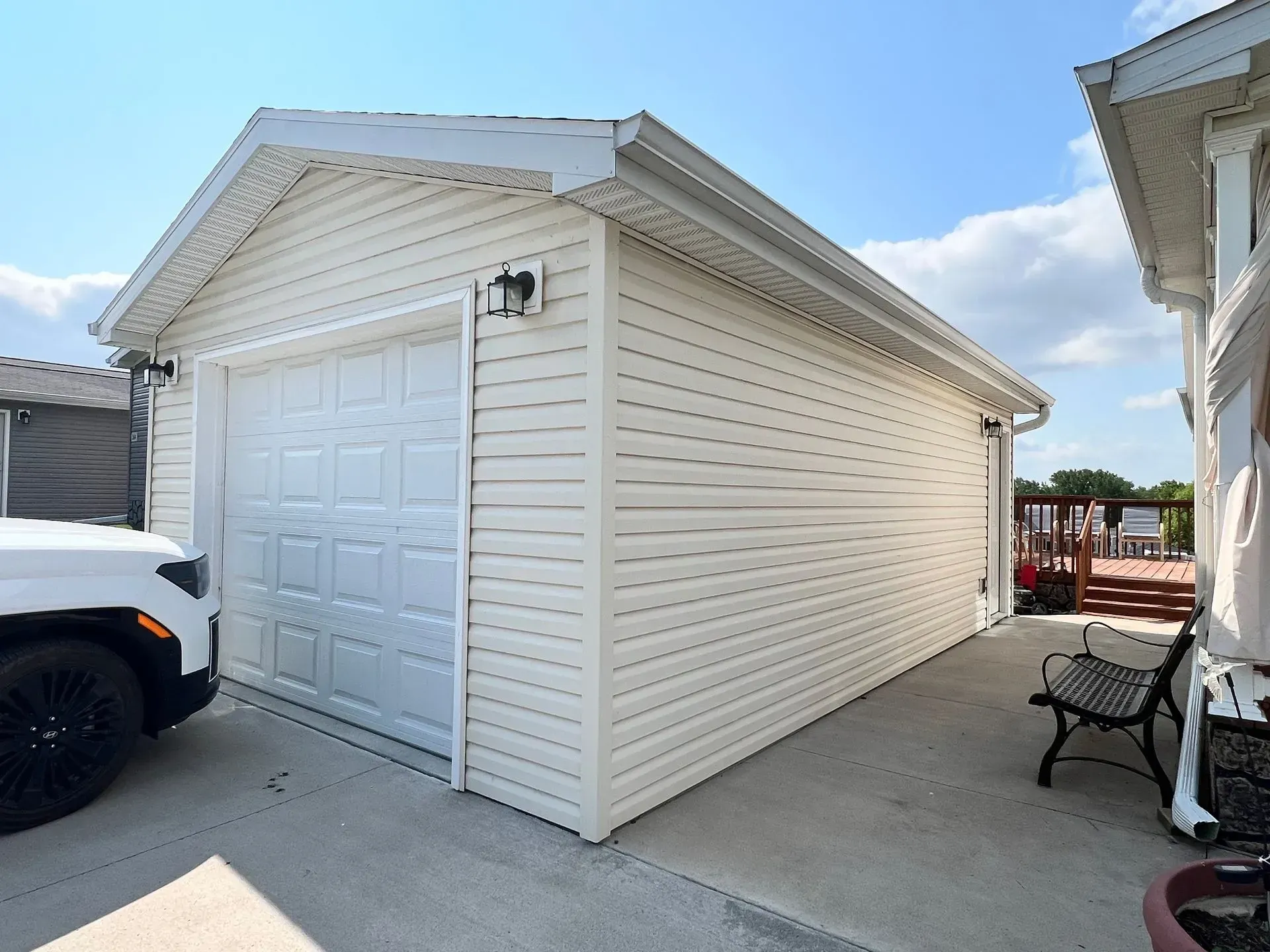 Tan vinyl-sided garage with white door and trim. A car is parked to the side, and a porch is visible behind it.