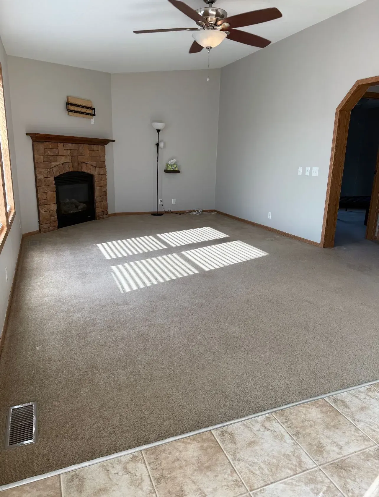 Spacious living room with a brick fireplace, beige carpet, and arched doorway. Sunlight streams in.