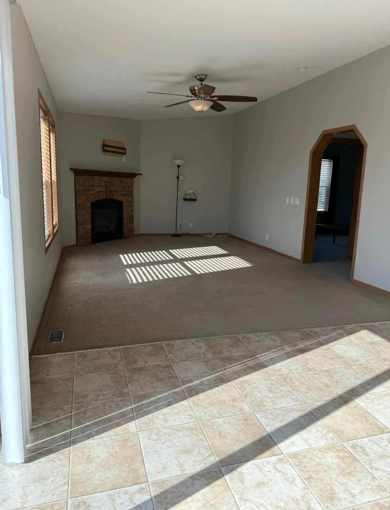 Empty living room with fireplace, ceiling fan, and arched doorway. Sunlight streams through windows.