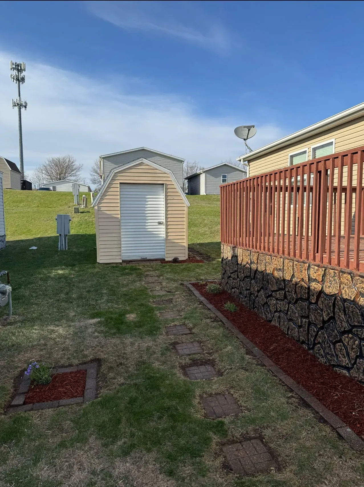 Backyard scene: a small storage shed, stepping stones, wooden deck with red railing, and blue sky.