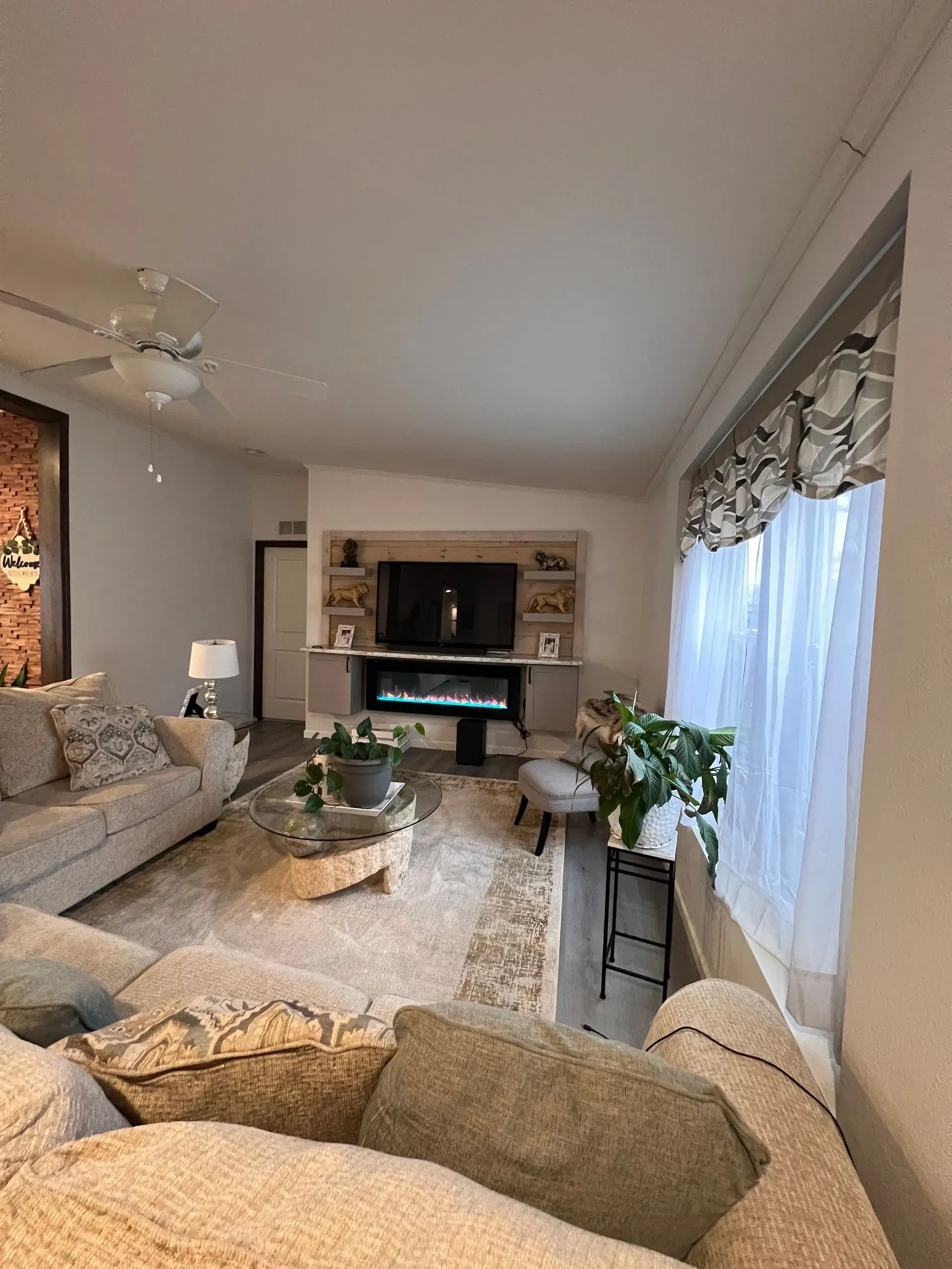 Cozy living room with a large TV, fireplace, and neutral-toned furniture. A window with sheer curtains is on the right.