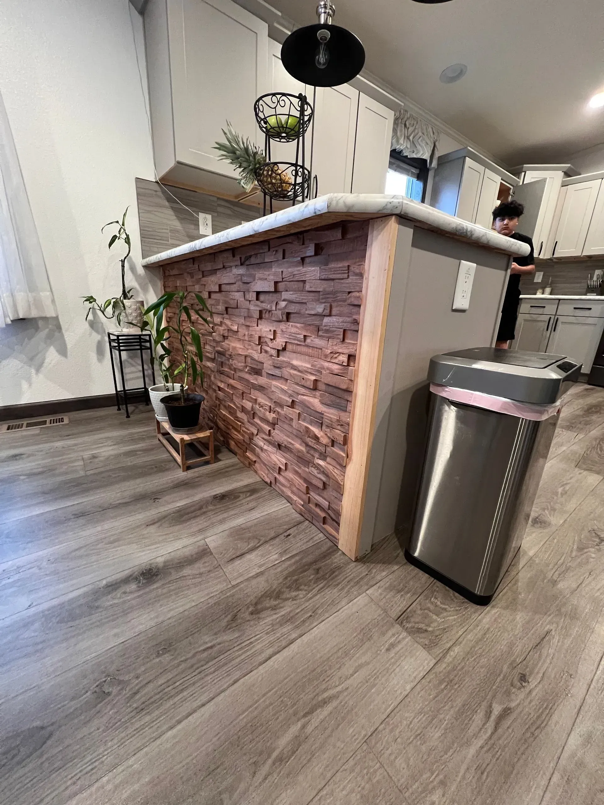 Kitchen island with brick-like facade, light wood trim, grey countertop. Stainless steel trash can. Grey wood flooring.