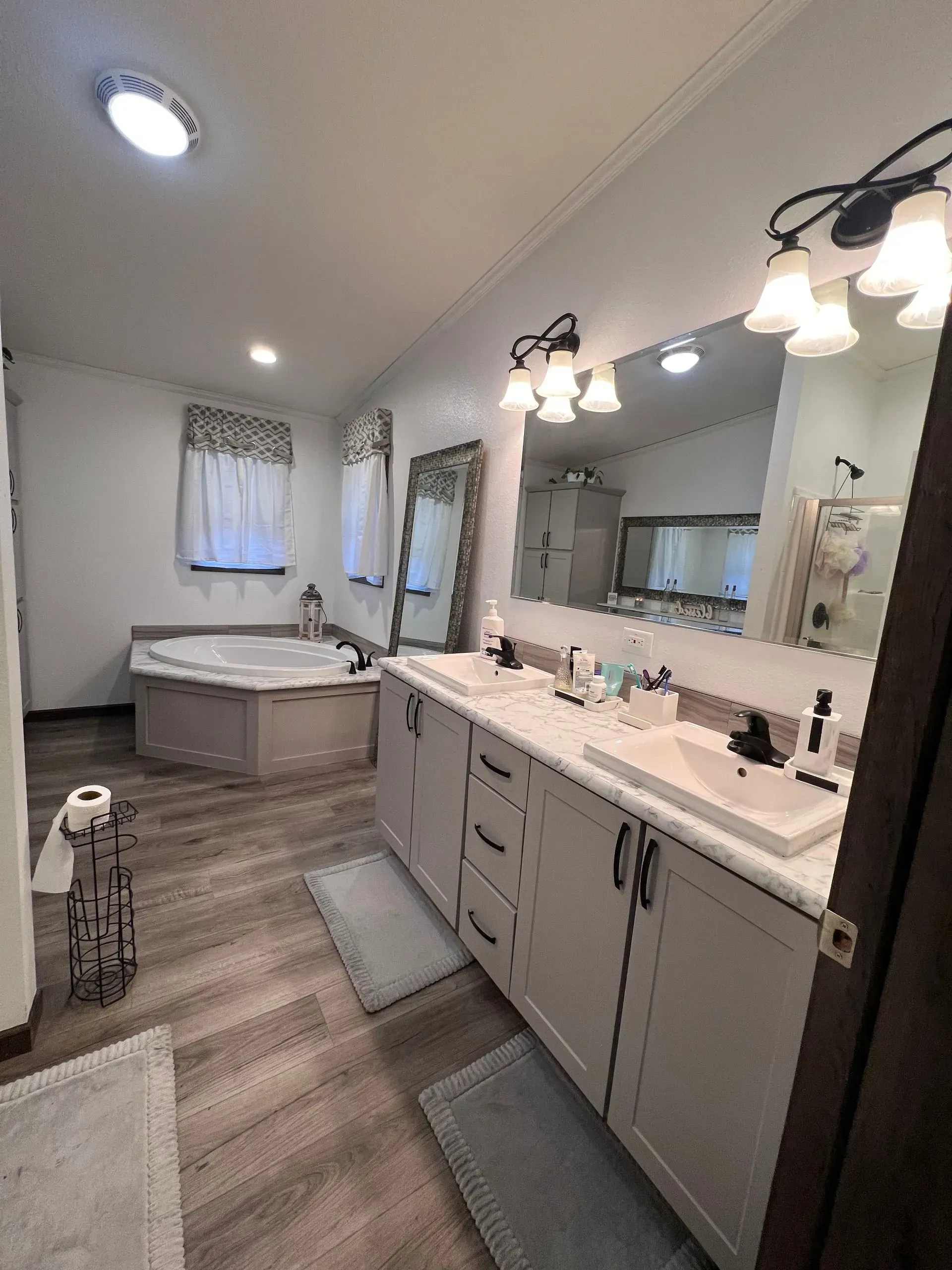 Bathroom with double vanity, bathtub, and large mirror. Light gray cabinets, wood-look floor, white walls.