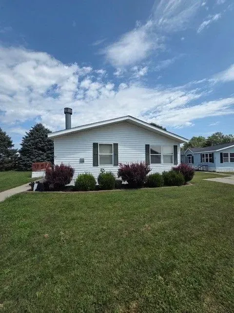White single-story house with green shutters, chimney, and bushes, set on a grassy lawn under a blue sky.