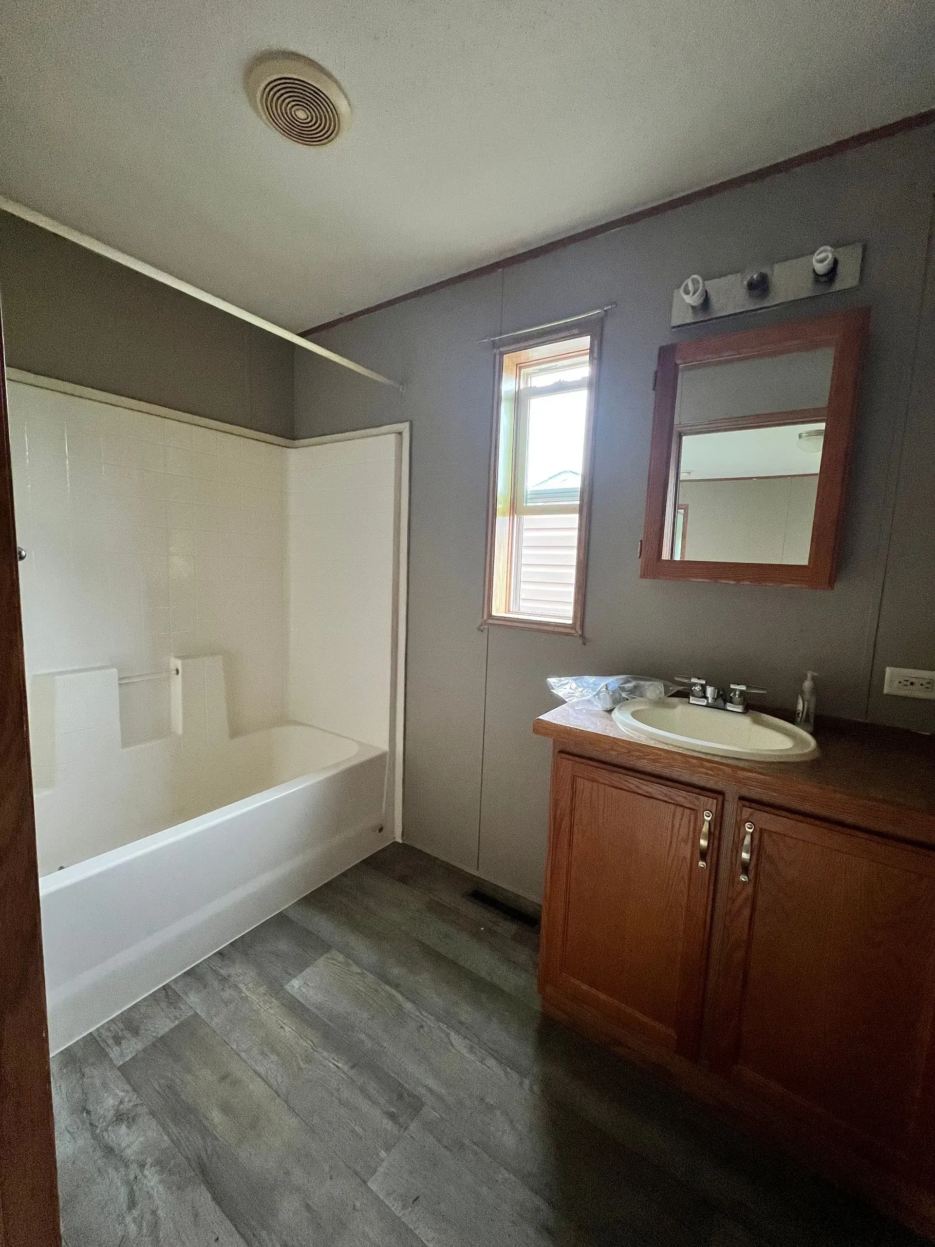 Bathroom with tub, vanity, mirror, window. Gray walls, brown cabinets, white tub.