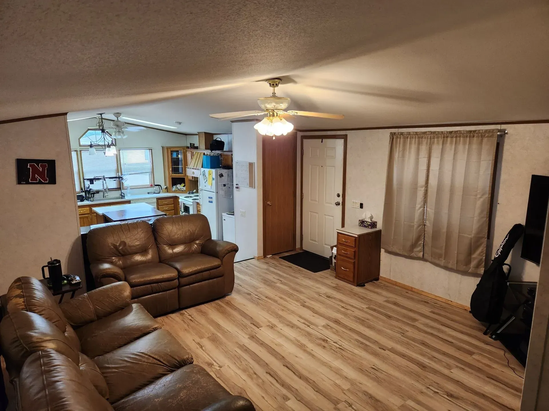 Living room with brown leather sofa, open to kitchen, door, and wood-look flooring.