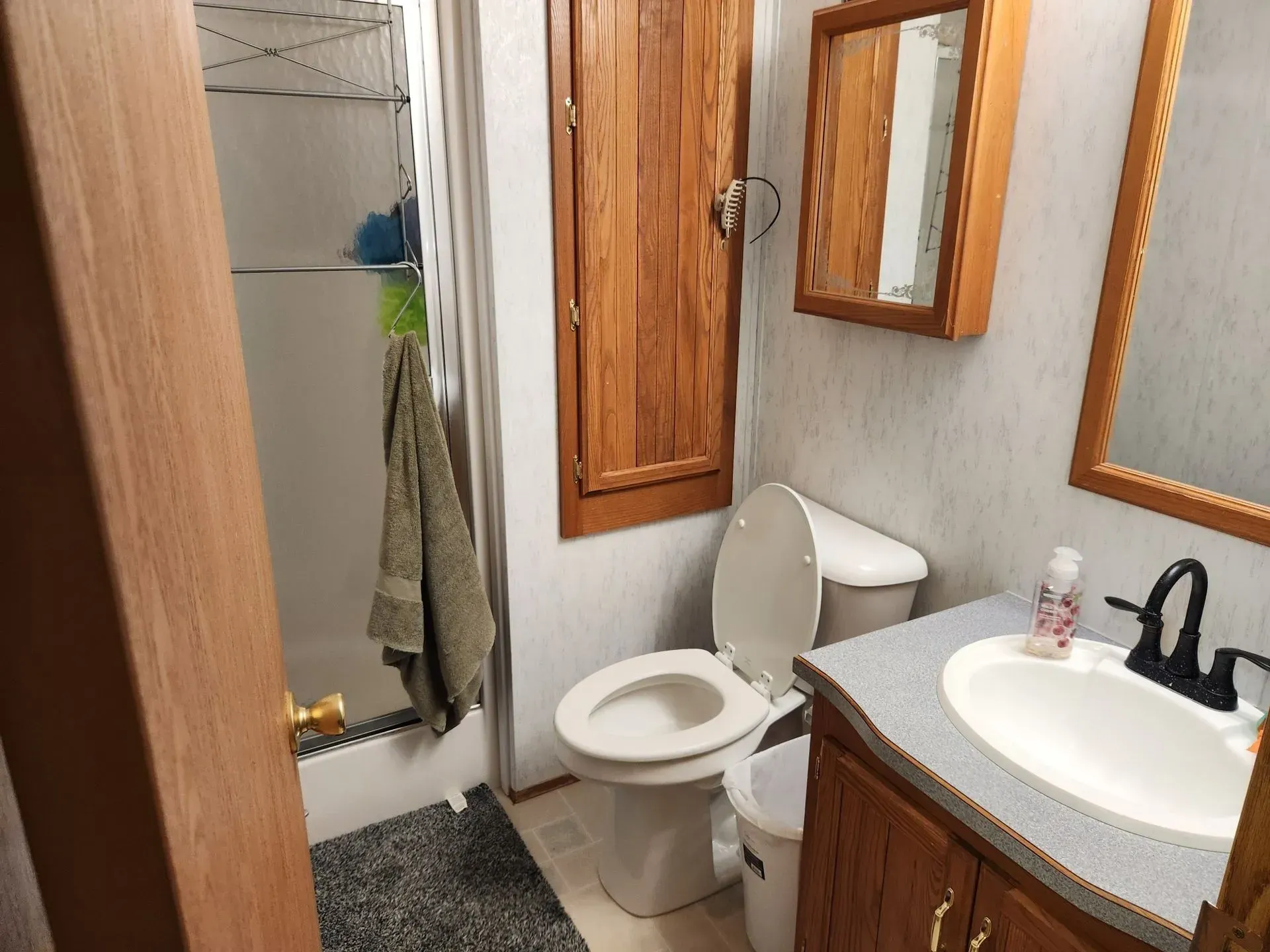 Bathroom with a toilet, sink, mirror, and shower, all in light colors and wood accents.