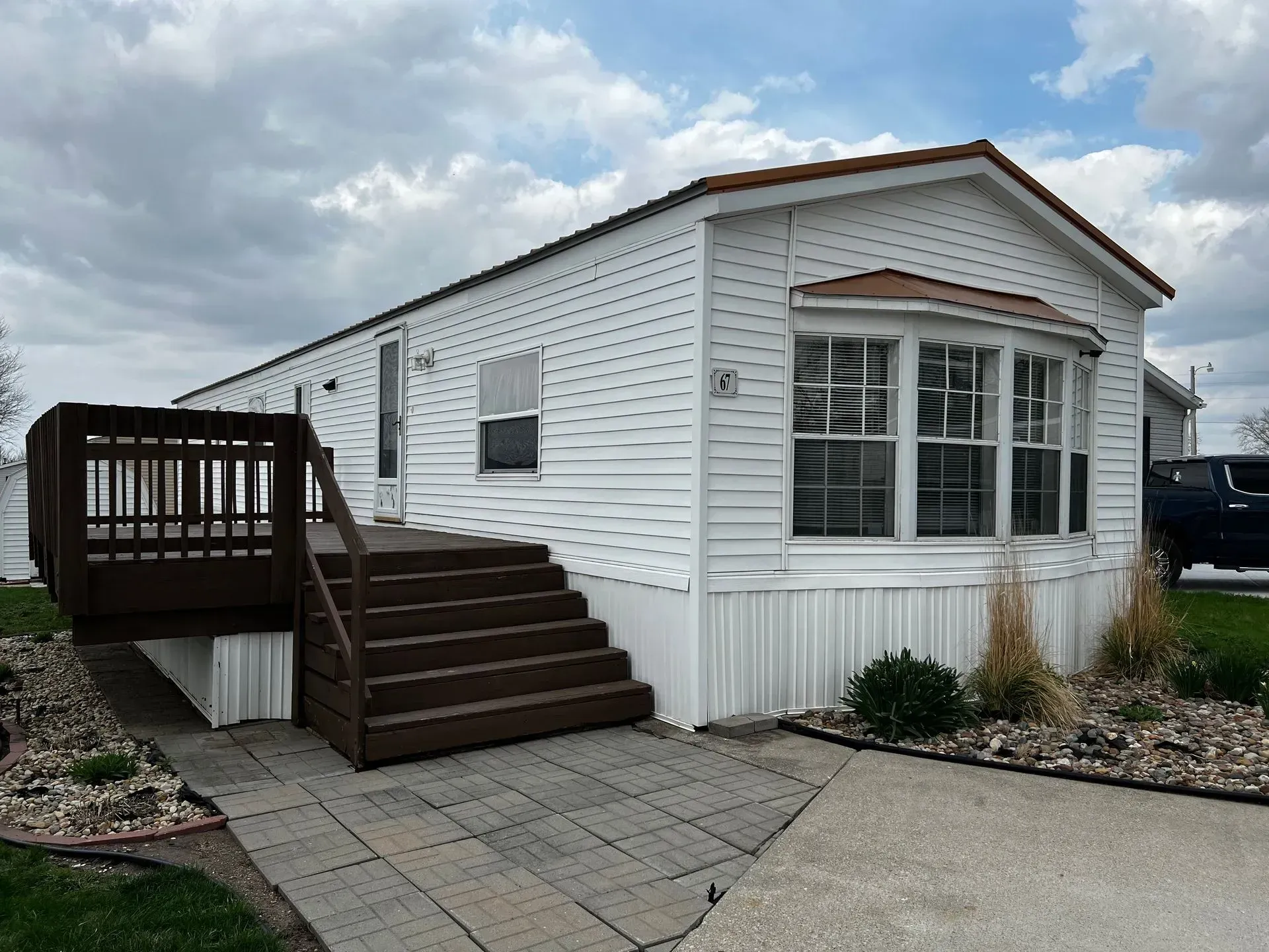 White mobile home with brown deck and stairs.  Paved walkway and cloudy sky.
