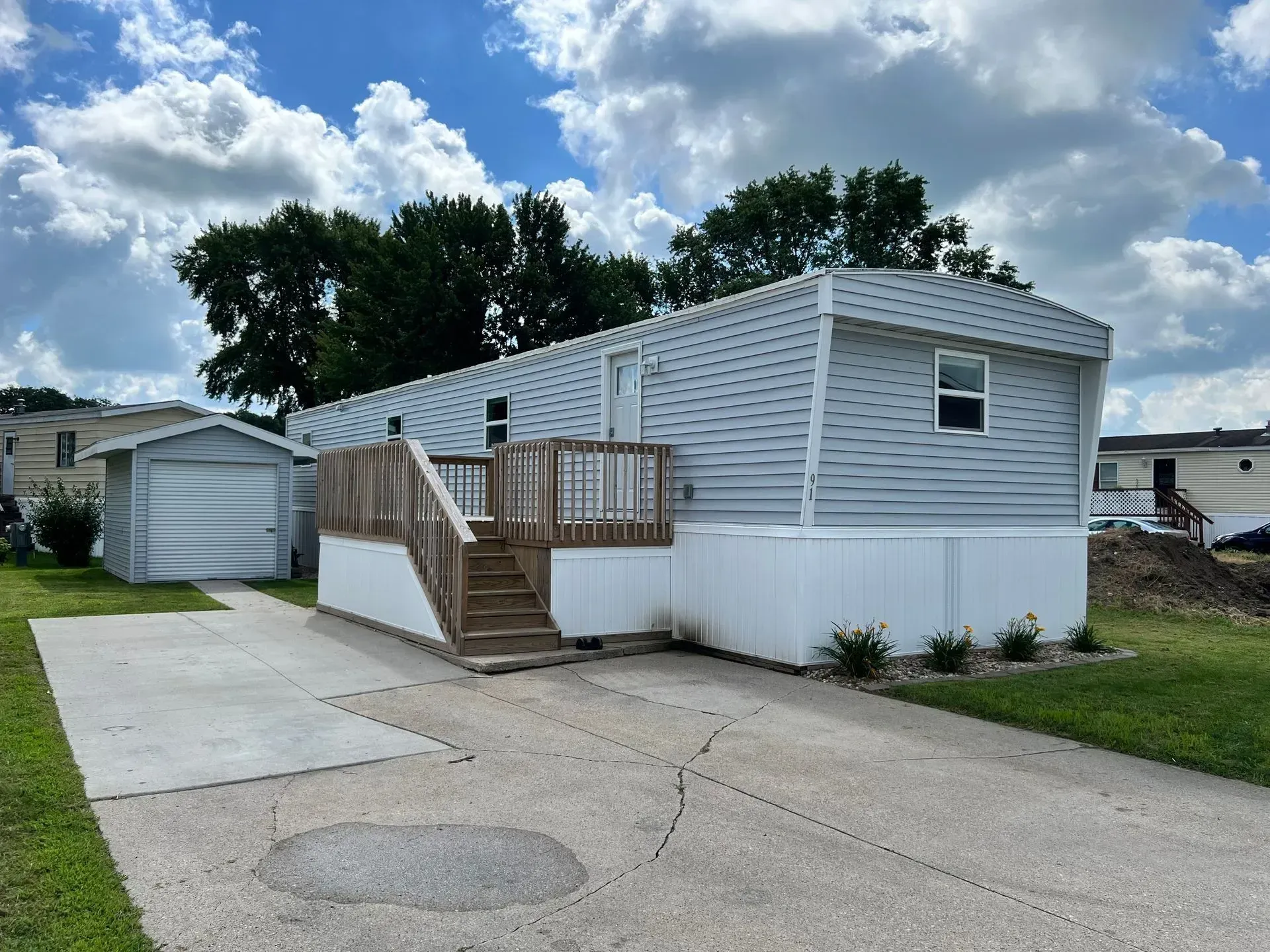 Mobile home with gray siding, wooden deck, and a detached garage, parked on a concrete driveway.