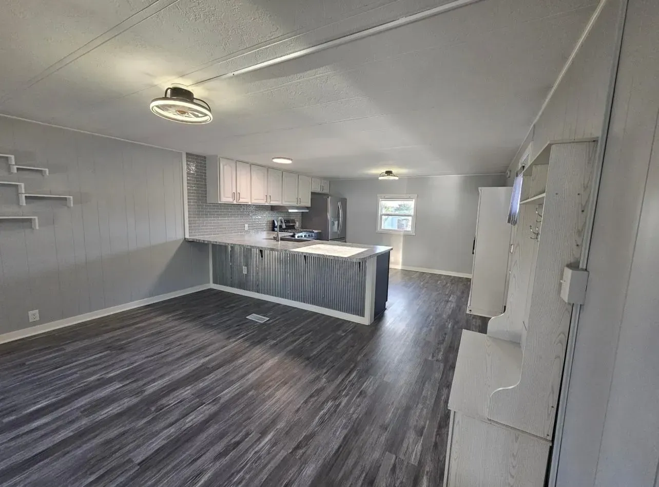 Open-concept kitchen and living area with gray flooring, cabinets, and island. Natural light from a window.